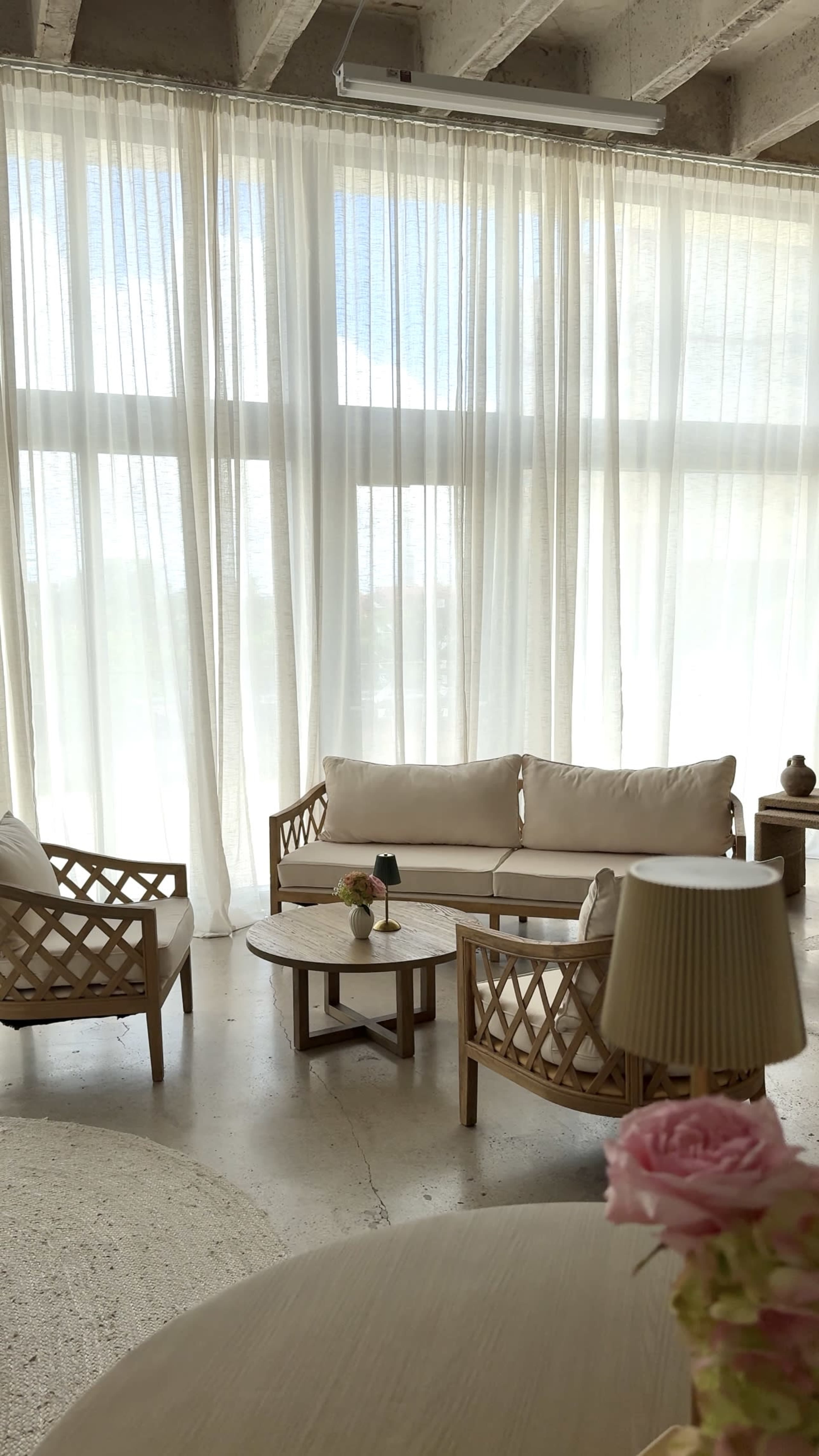 A sunlit room features a light-colored sofa and two matching chairs arranged around a round table, with sheer curtains filtering the light from large windows.