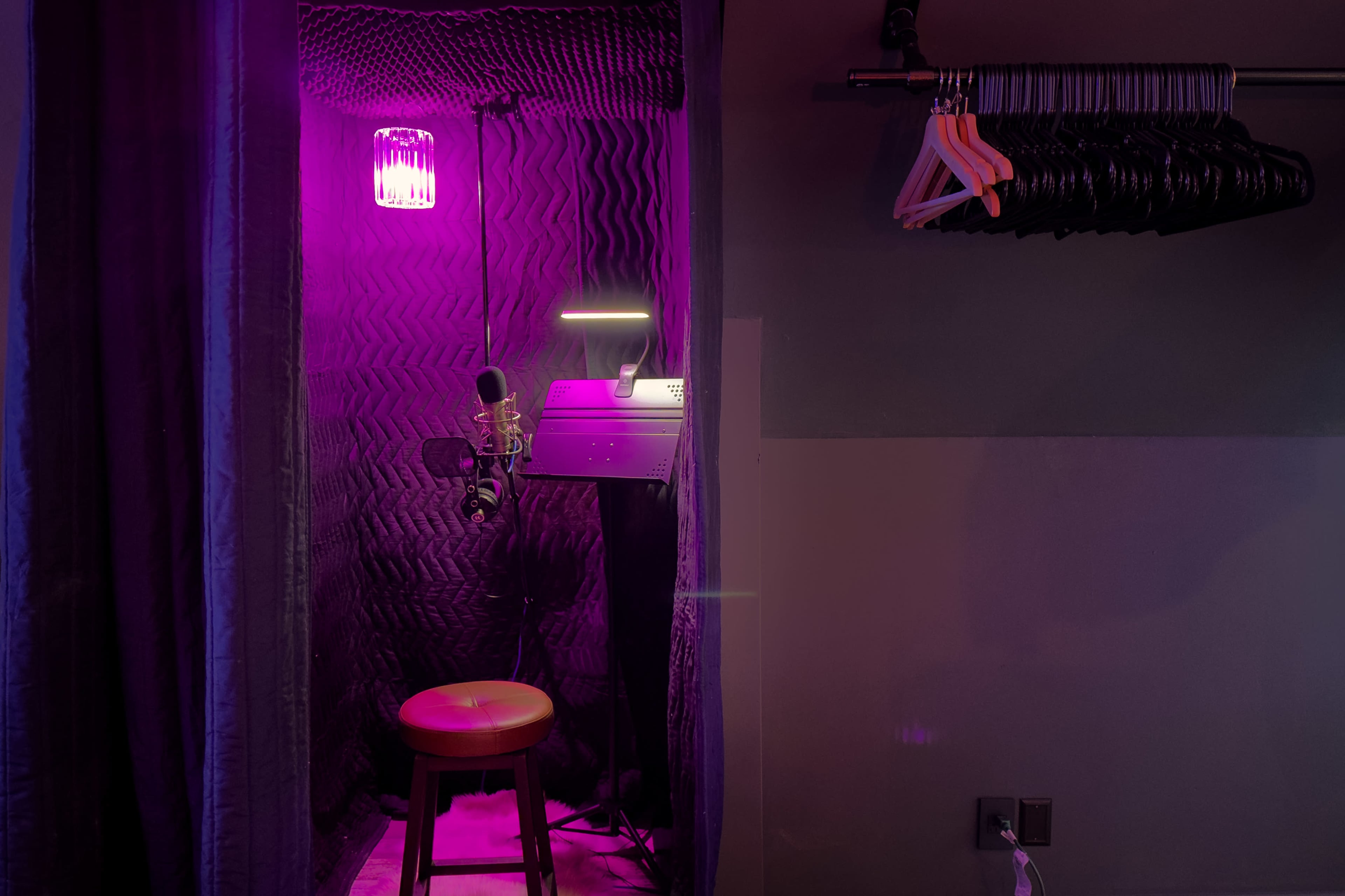 A small soundproof booth features a microphone, a stool, and purple lighting, with hangers and a wall on the side.