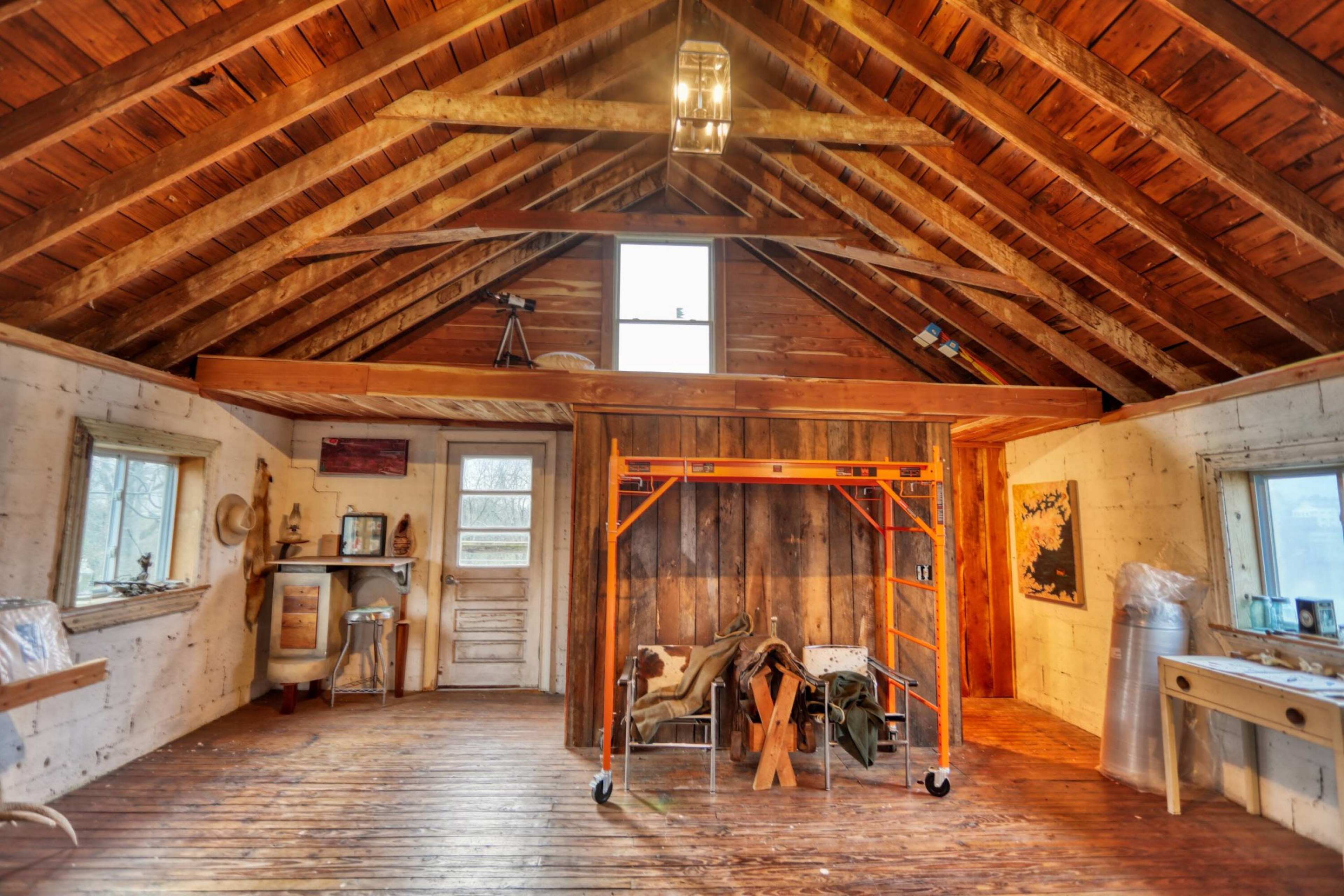 The interior of a rustic cabin with wooden beams, a wooden floor, and various tools and furnishings scattered around.
