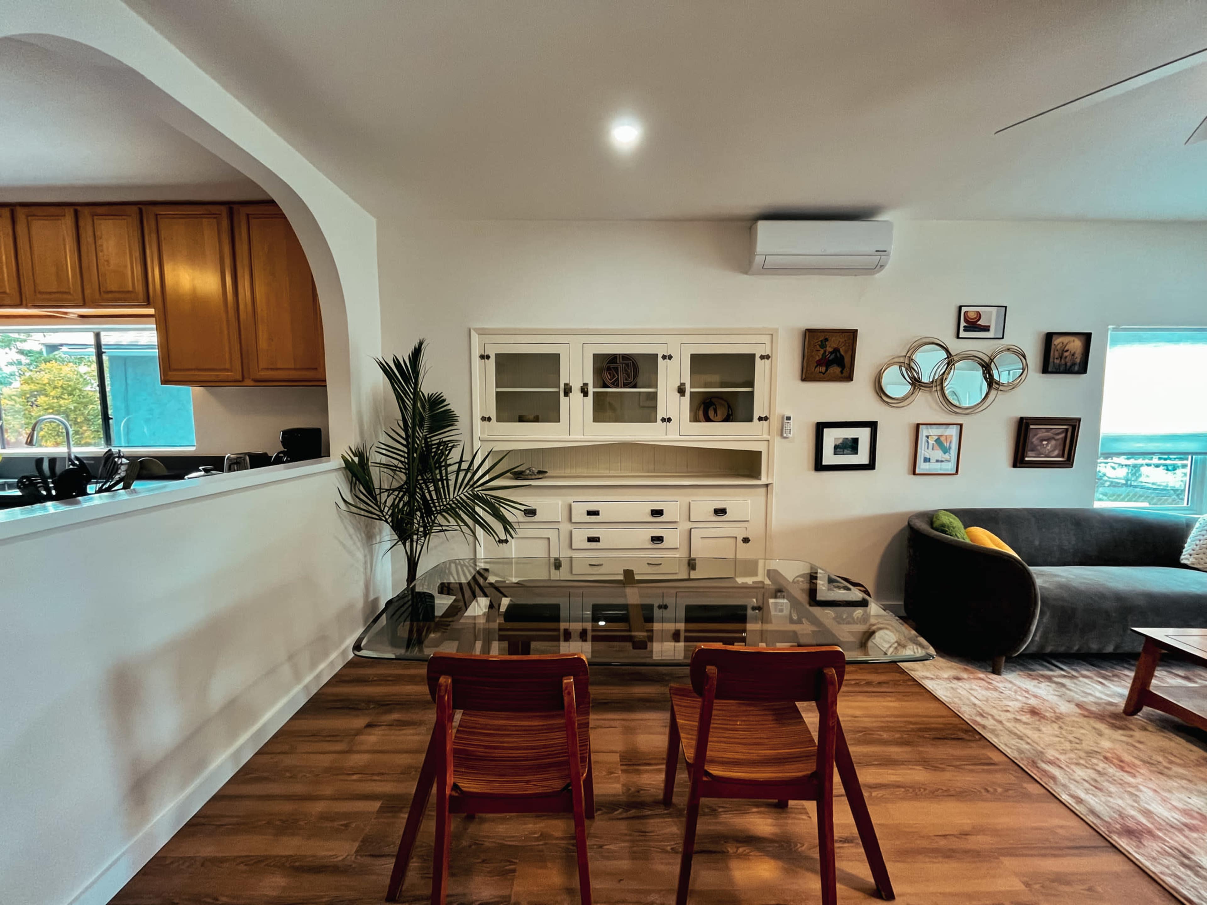 A modern living space features a glass dining table with two red chairs, a dark sofa, and wall decor, all set against a backdrop of wooden cabinetry and a kitchen area.