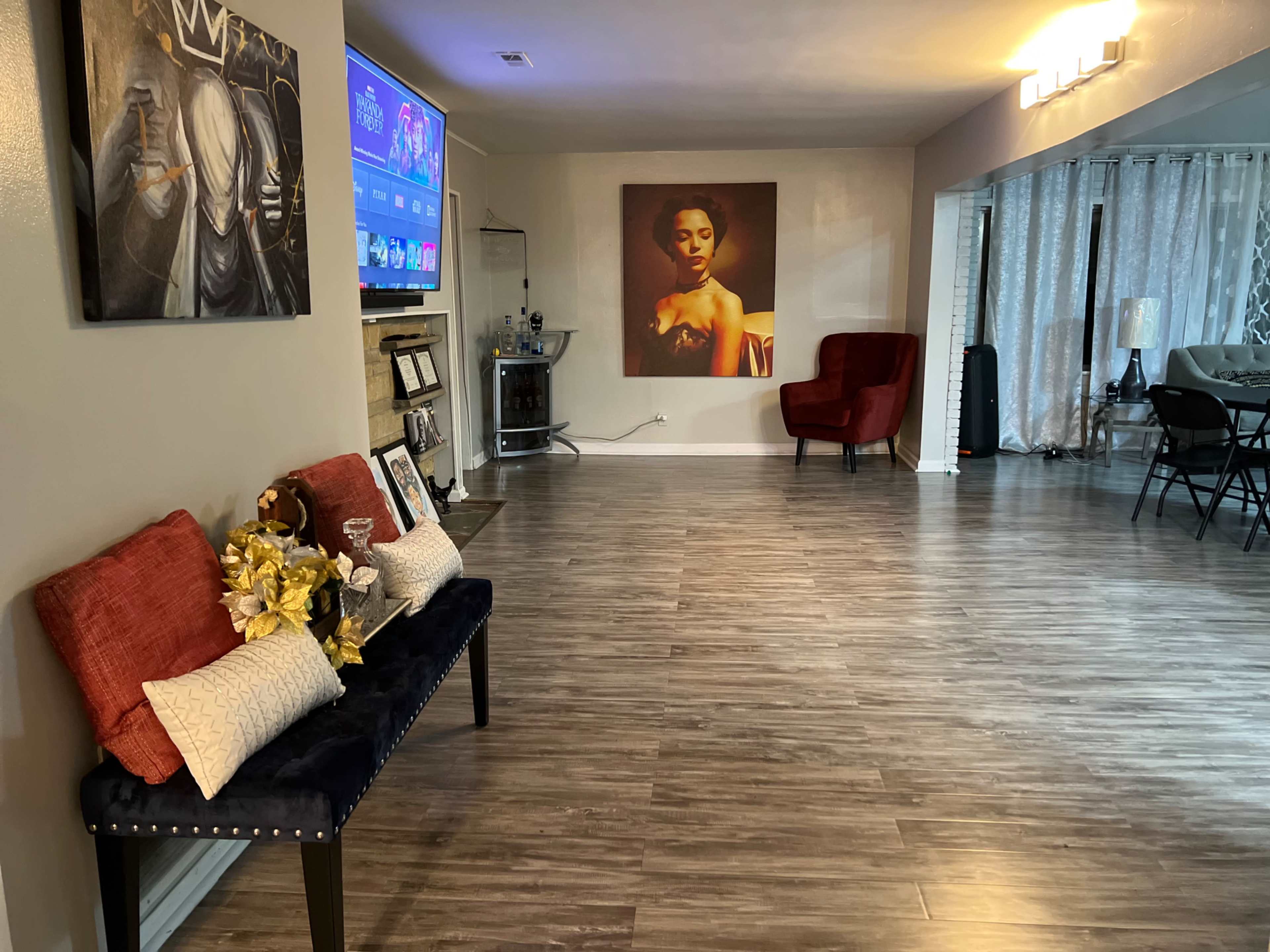 The image shows a spacious living area featuring a seating arrangement with decorative pillows, a television on the wall, and artwork on the adjacent wall.