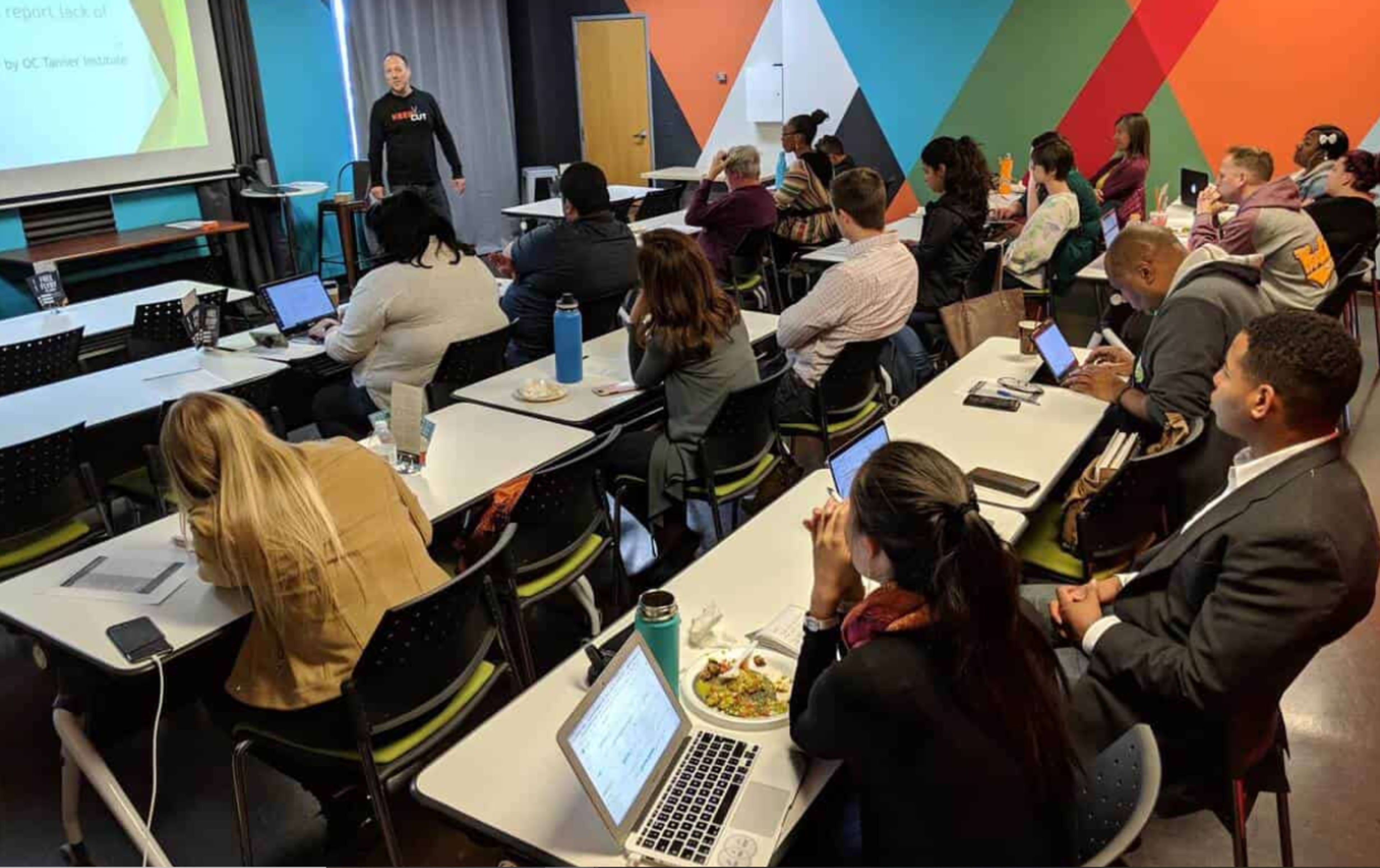 A presenter stands at the front of a classroom while an audience of seated participants listens attentively, with laptops and notebooks in front of them.