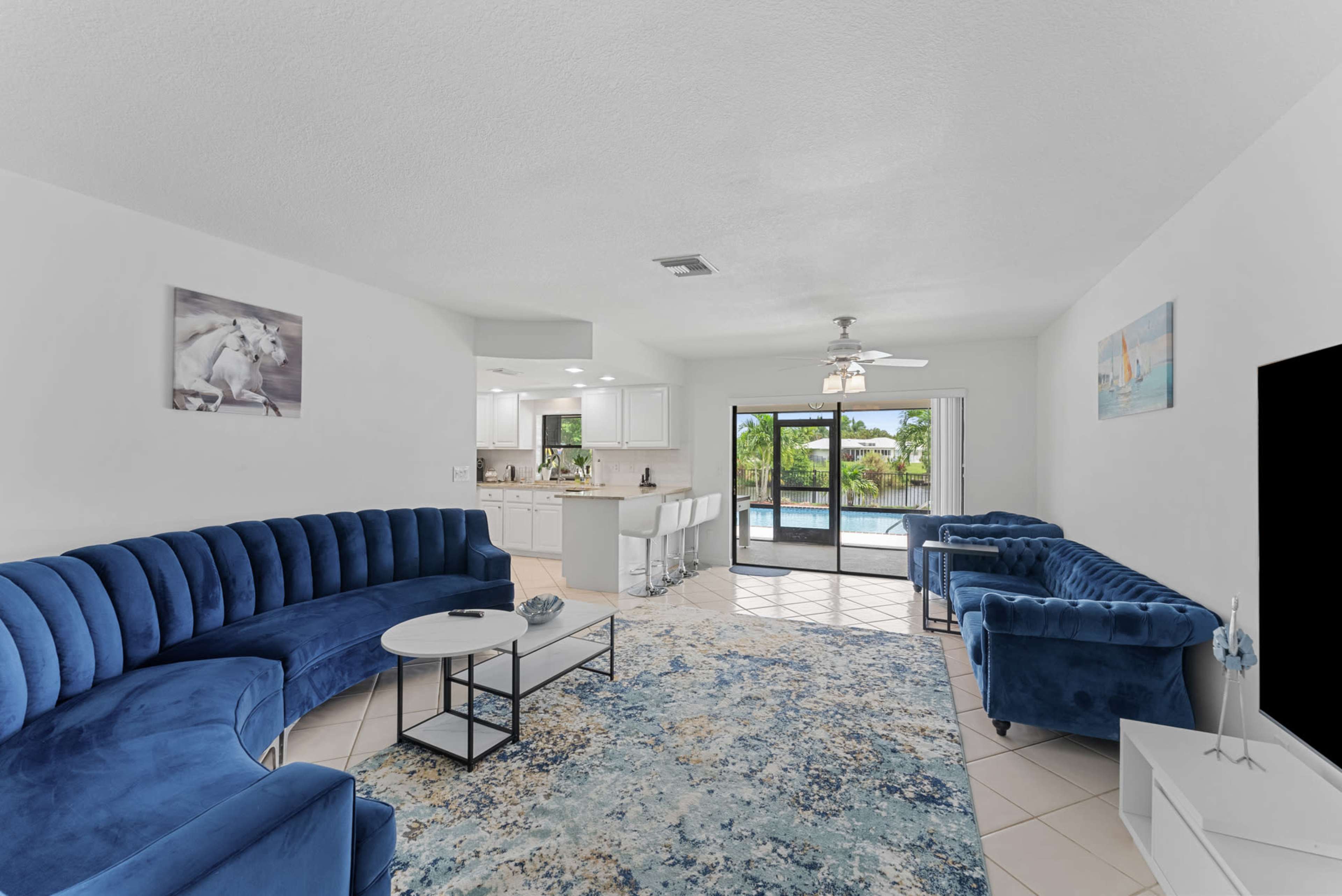 A bright living room features blue velvet sofas and a circular coffee table, opening up to a view of a pool area through sliding glass doors.