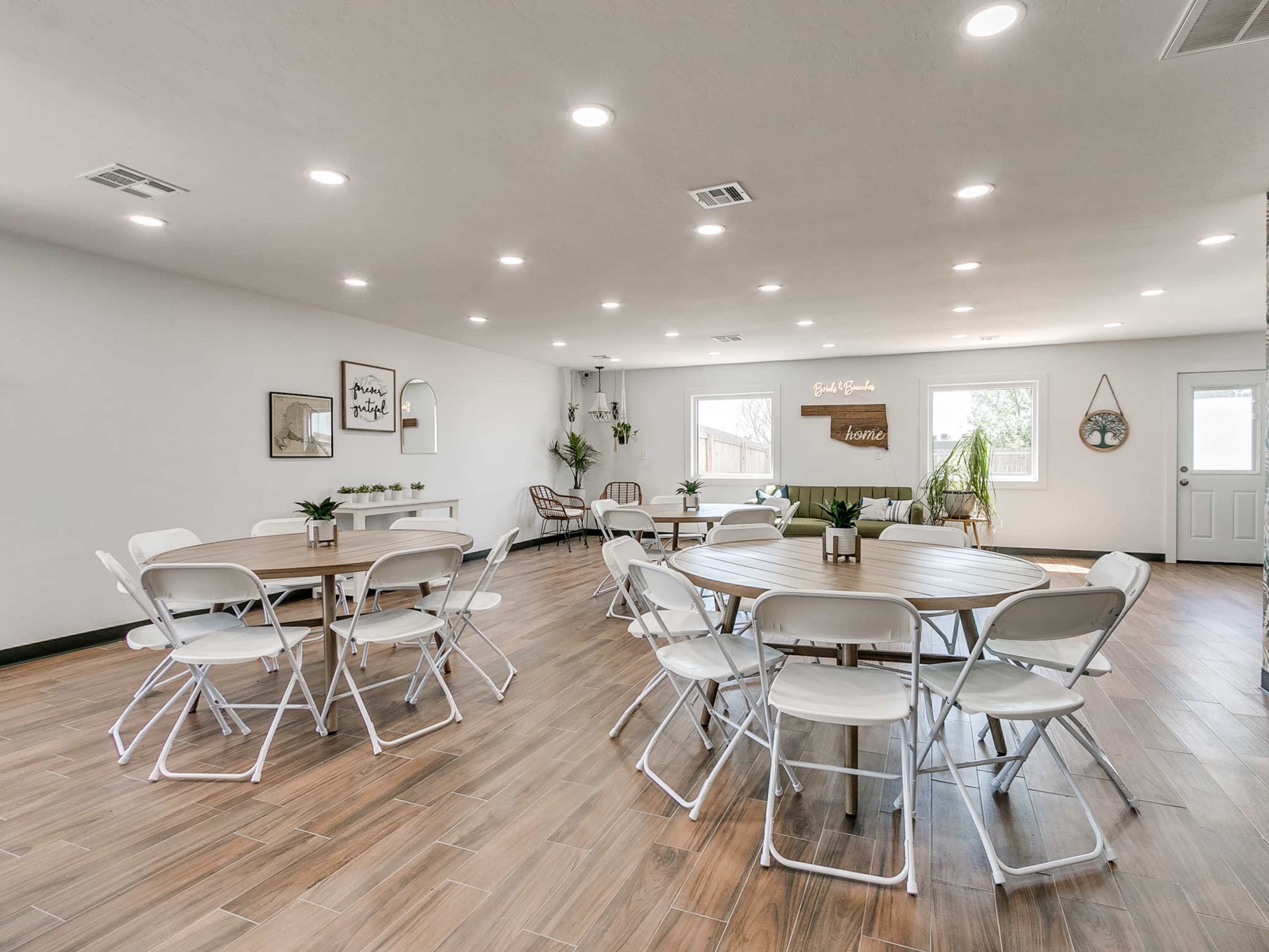 A spacious, well-lit room with multiple round tables and chairs arranged neatly, along with potted plants and a door leading outside.