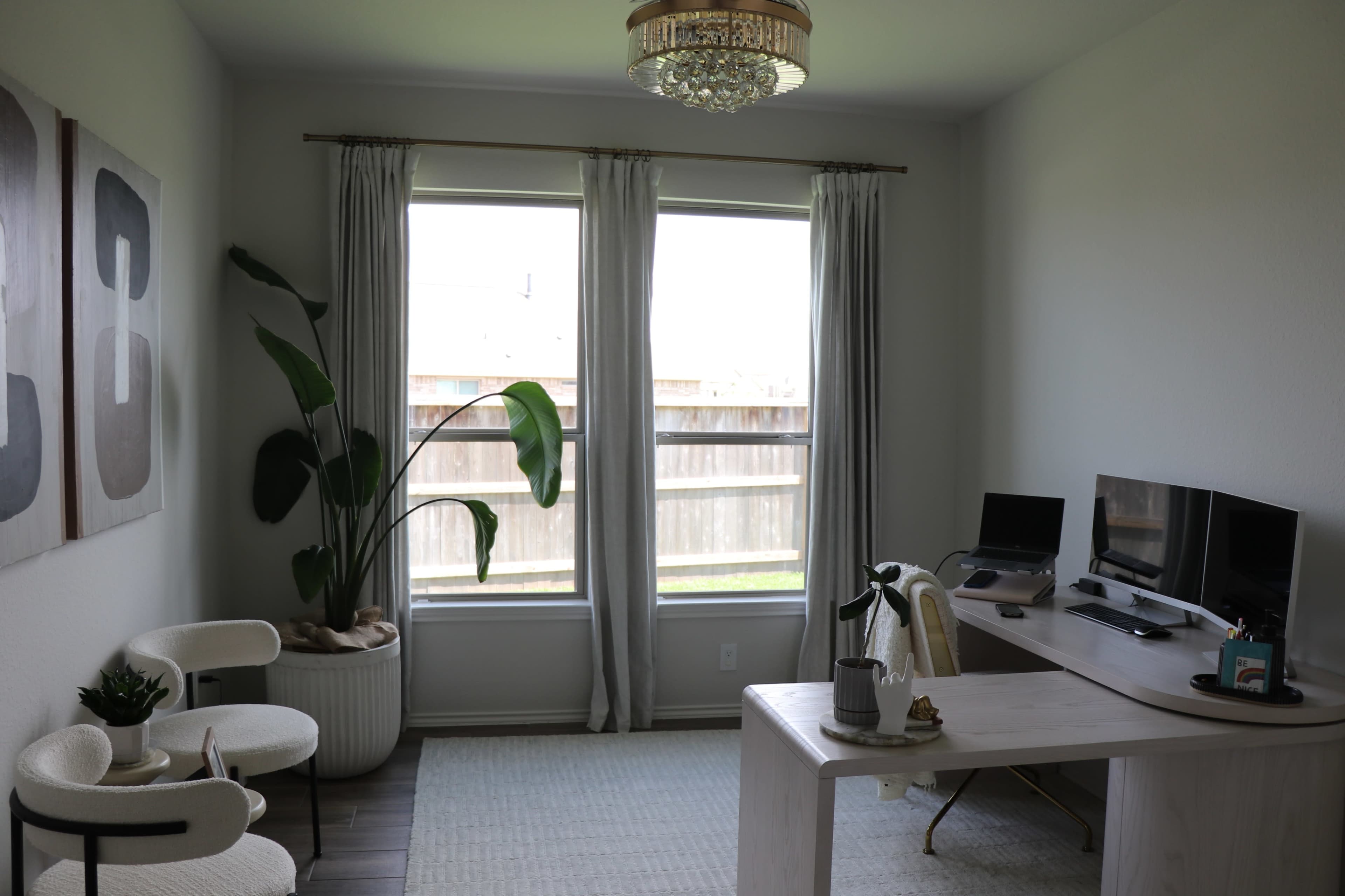 A home office features a desk with two computers, a large plant, and two chairs, illuminated by natural light from two windows.