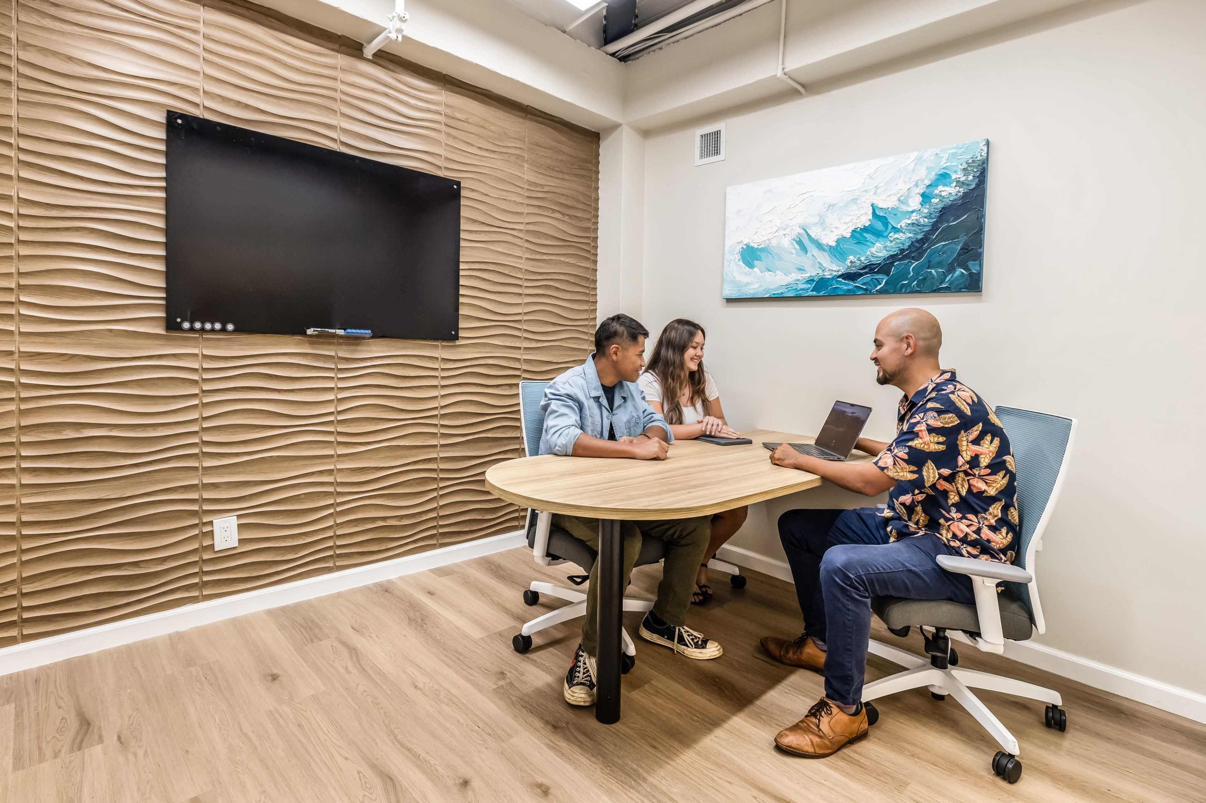 Three people are seated around a round table in a meeting room, with a wall-mounted screen in the background.