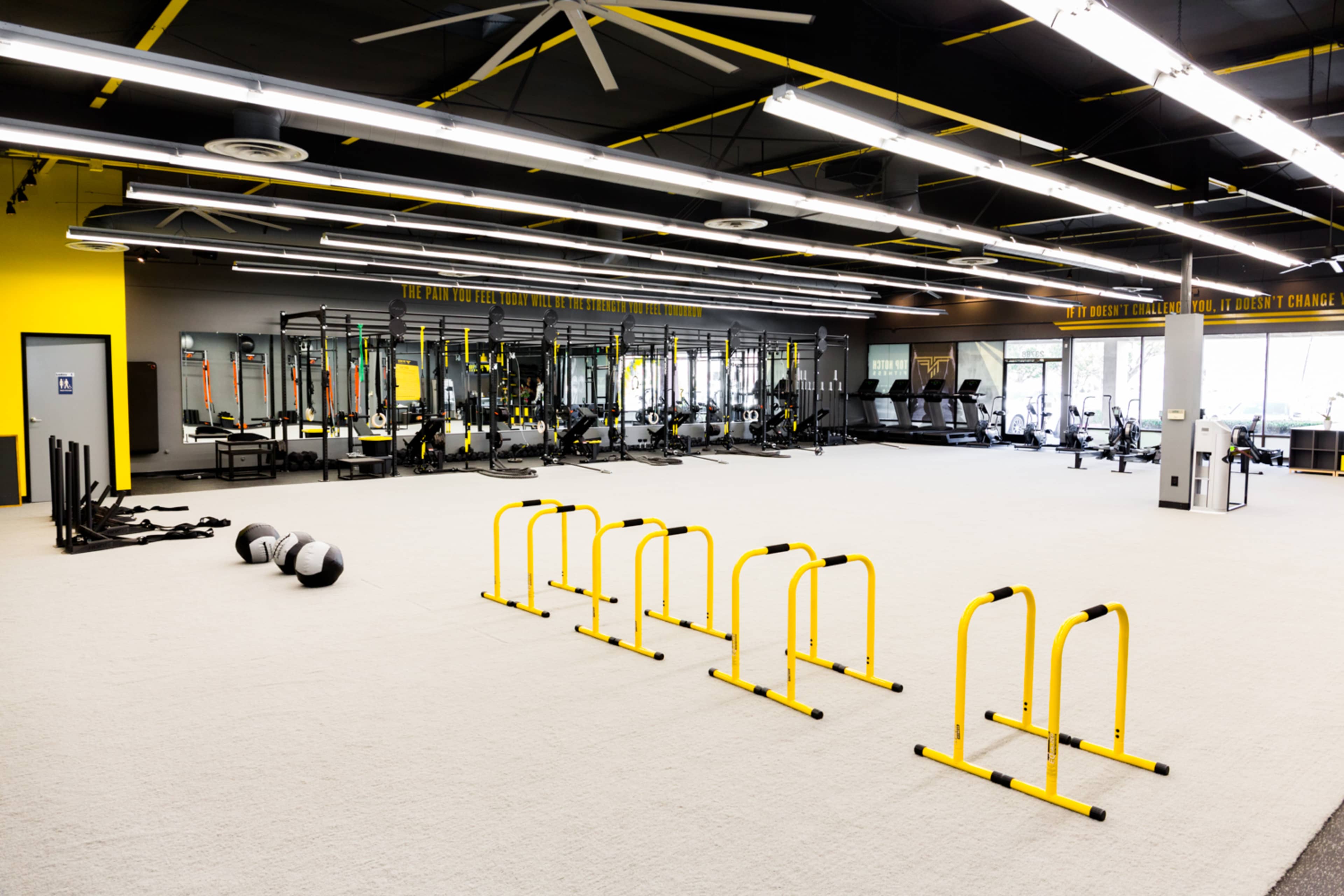 A spacious gym with various workout equipment, including treadmills, weight racks, and agility hurdles arranged on a light-colored floor.