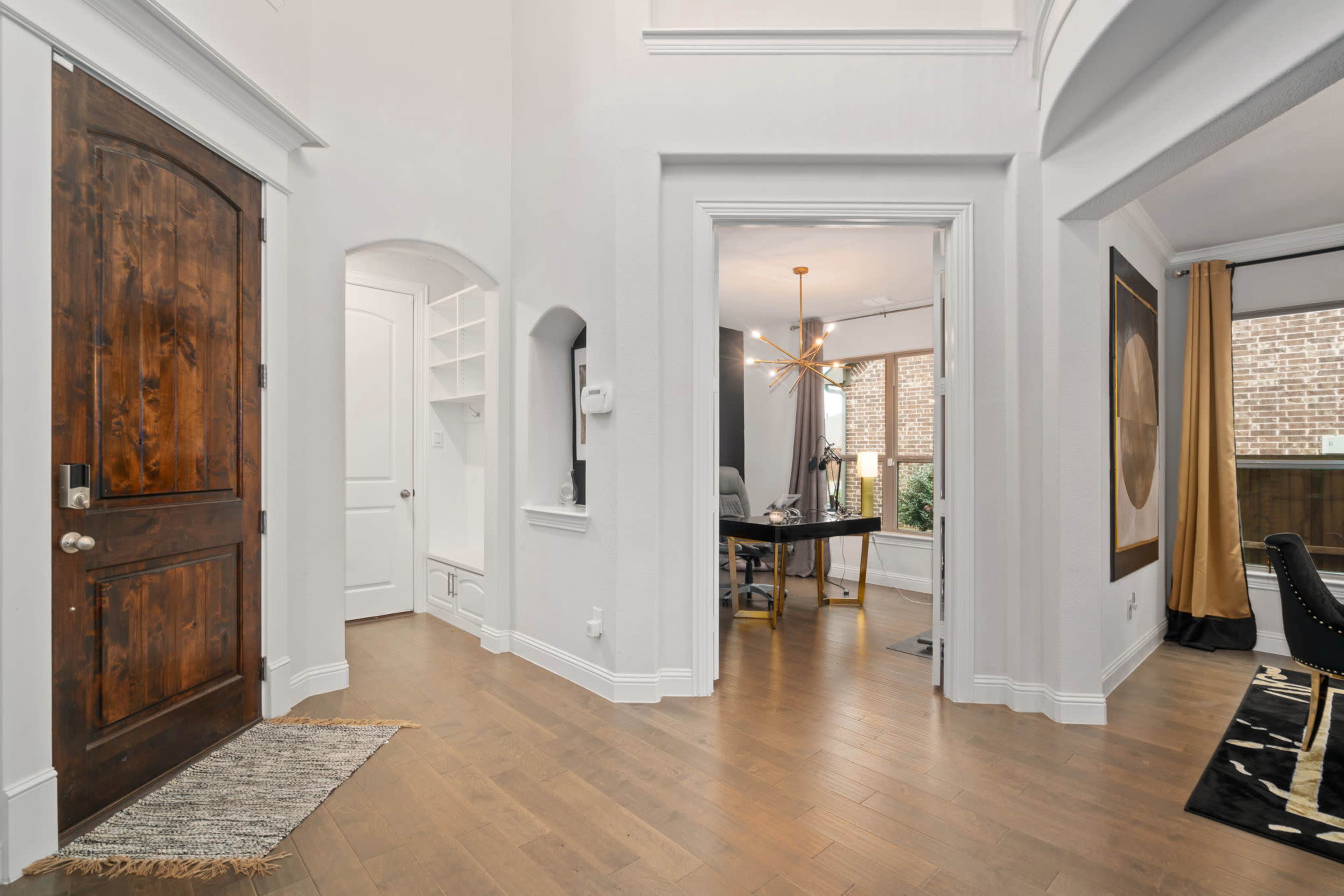 A spacious, well-lit foyer leading to a home office, with a wooden front door and light-colored walls.