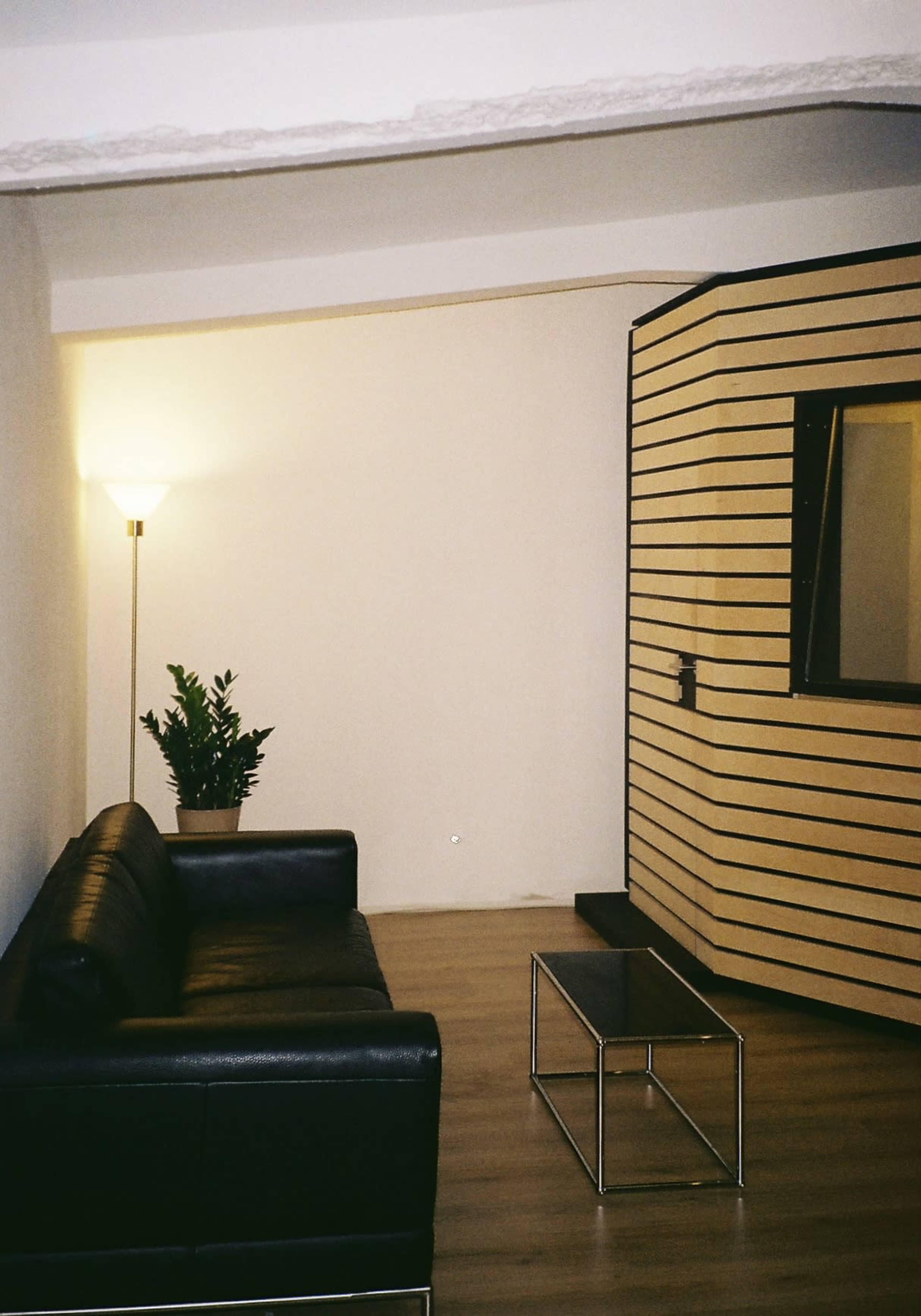 The image shows a modern interior space featuring a black leather sofa, a coffee table, and a partitioned area with striped walls and a mirror.