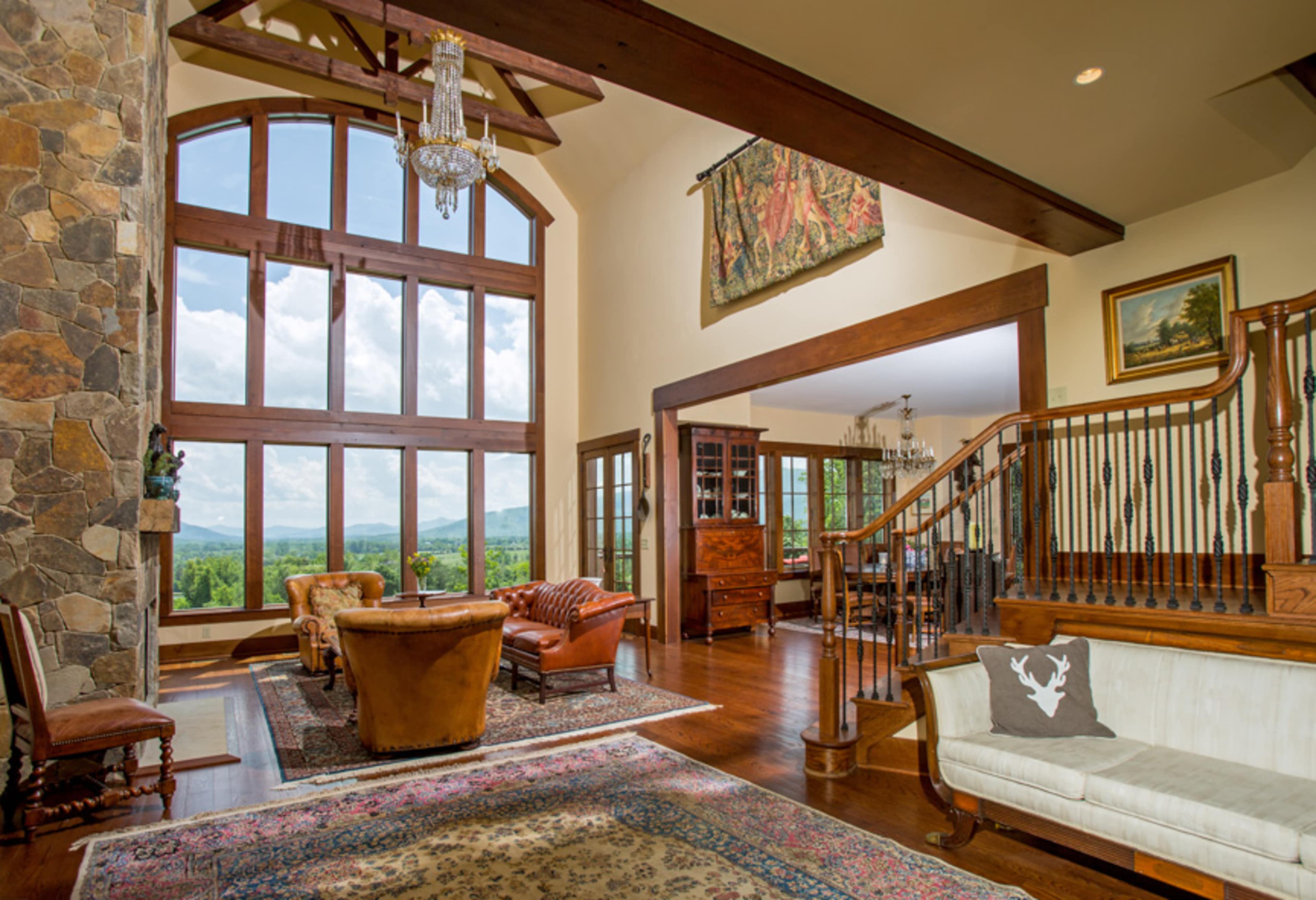 The spacious living room features large windows that frame a mountainous view, with a chandelier hanging above leather seating and a stone fireplace.