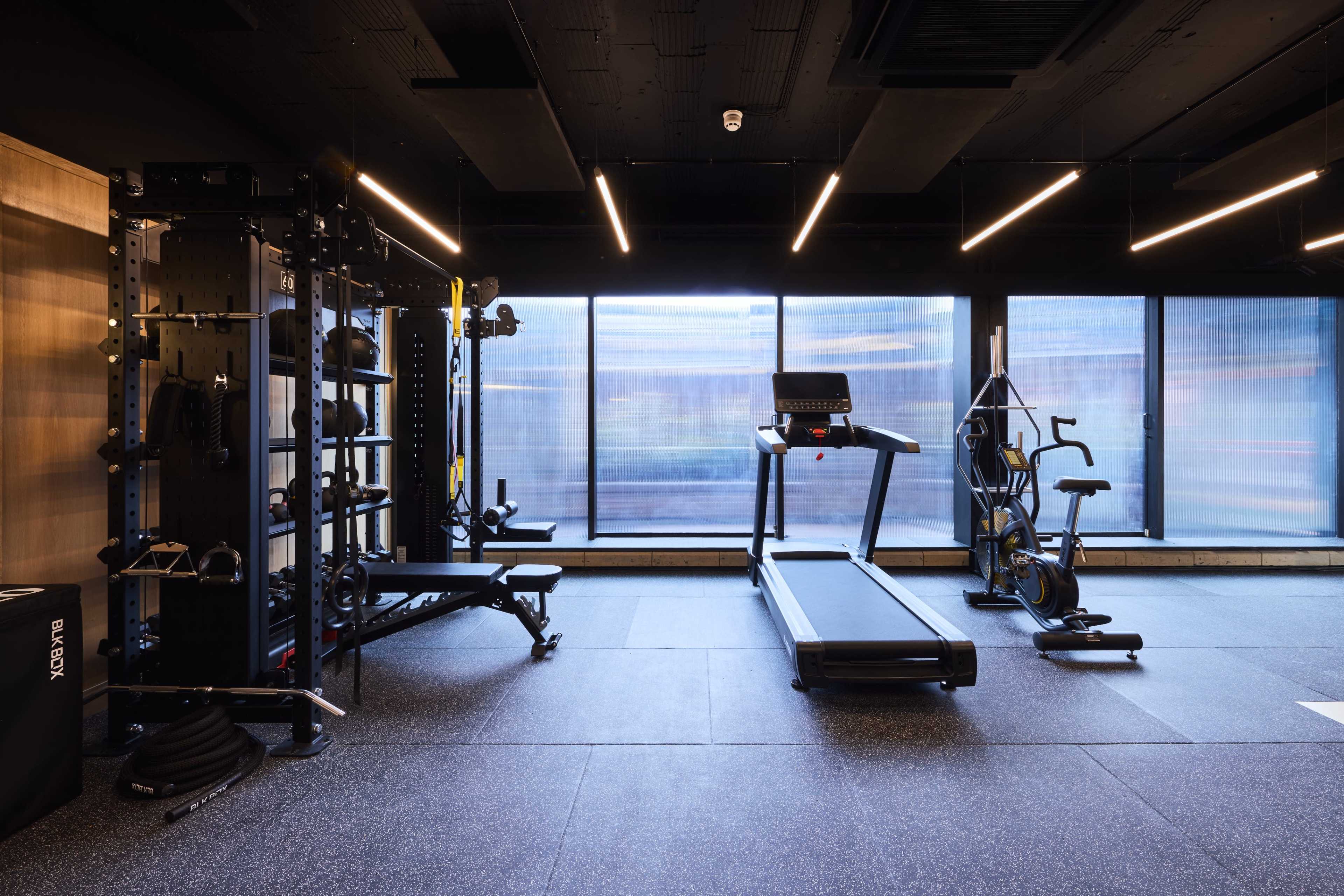 The image shows a modern gym interior featuring a treadmill, an exercise bike, a strength training station, and large windows allowing natural light to enter.