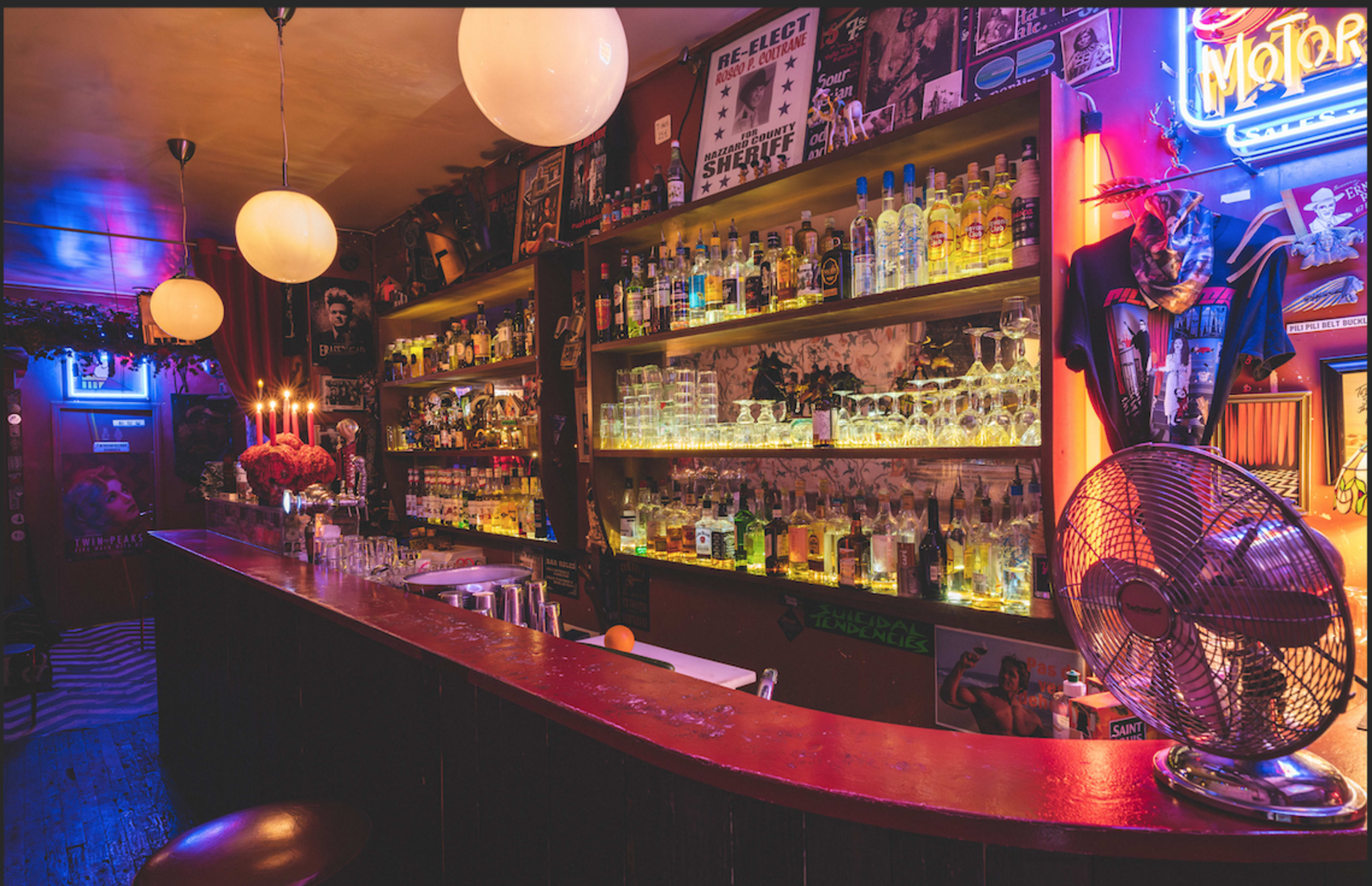 The image shows a dimly lit bar with a well-stocked back shelf of colorful liquor bottles and illuminated glasses, complemented by decorative elements and wall posters.