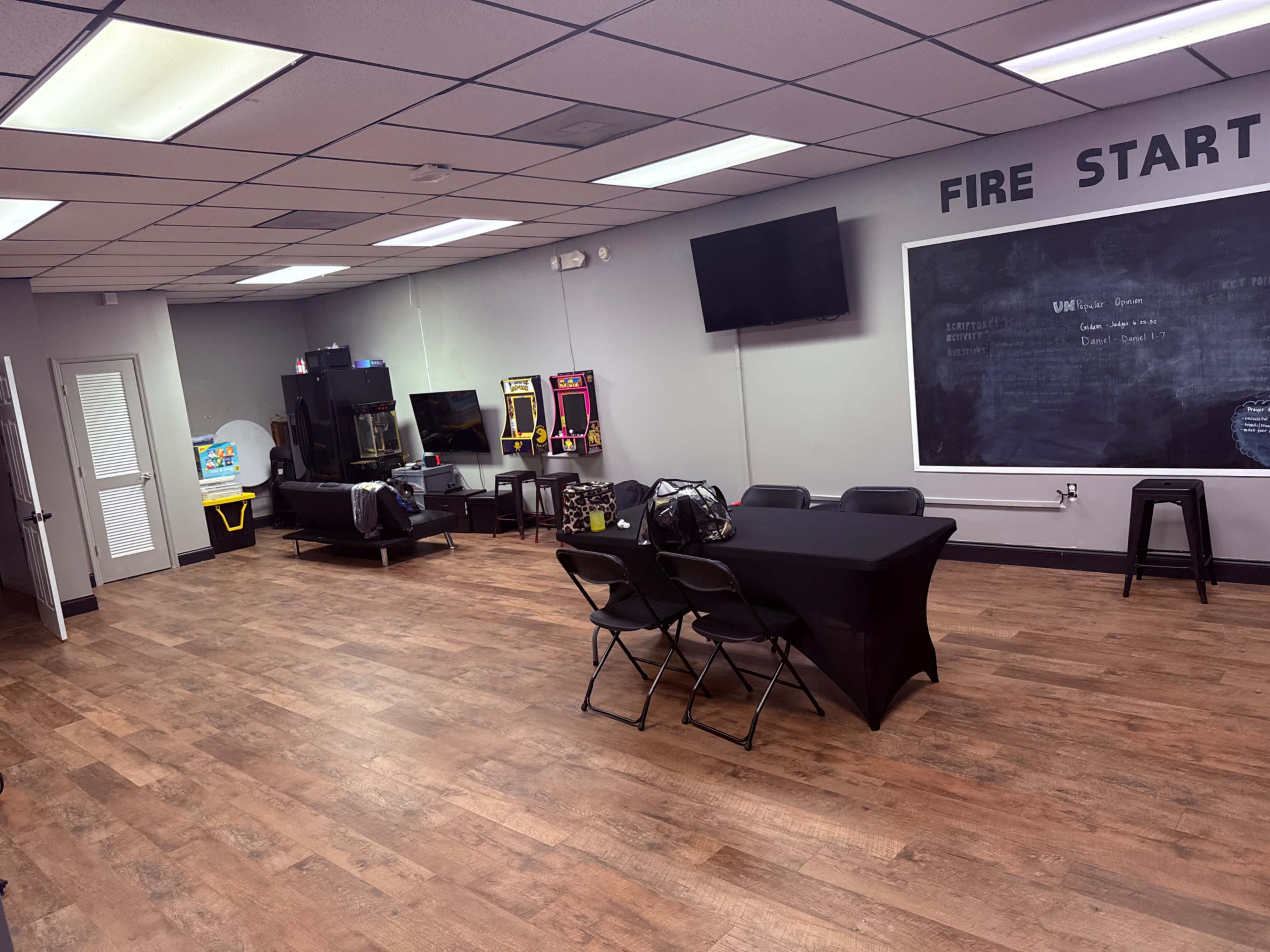 The image shows a spacious room featuring arcade machines, a black table with chairs, and a chalkboard wall, with wooden flooring and gray walls.