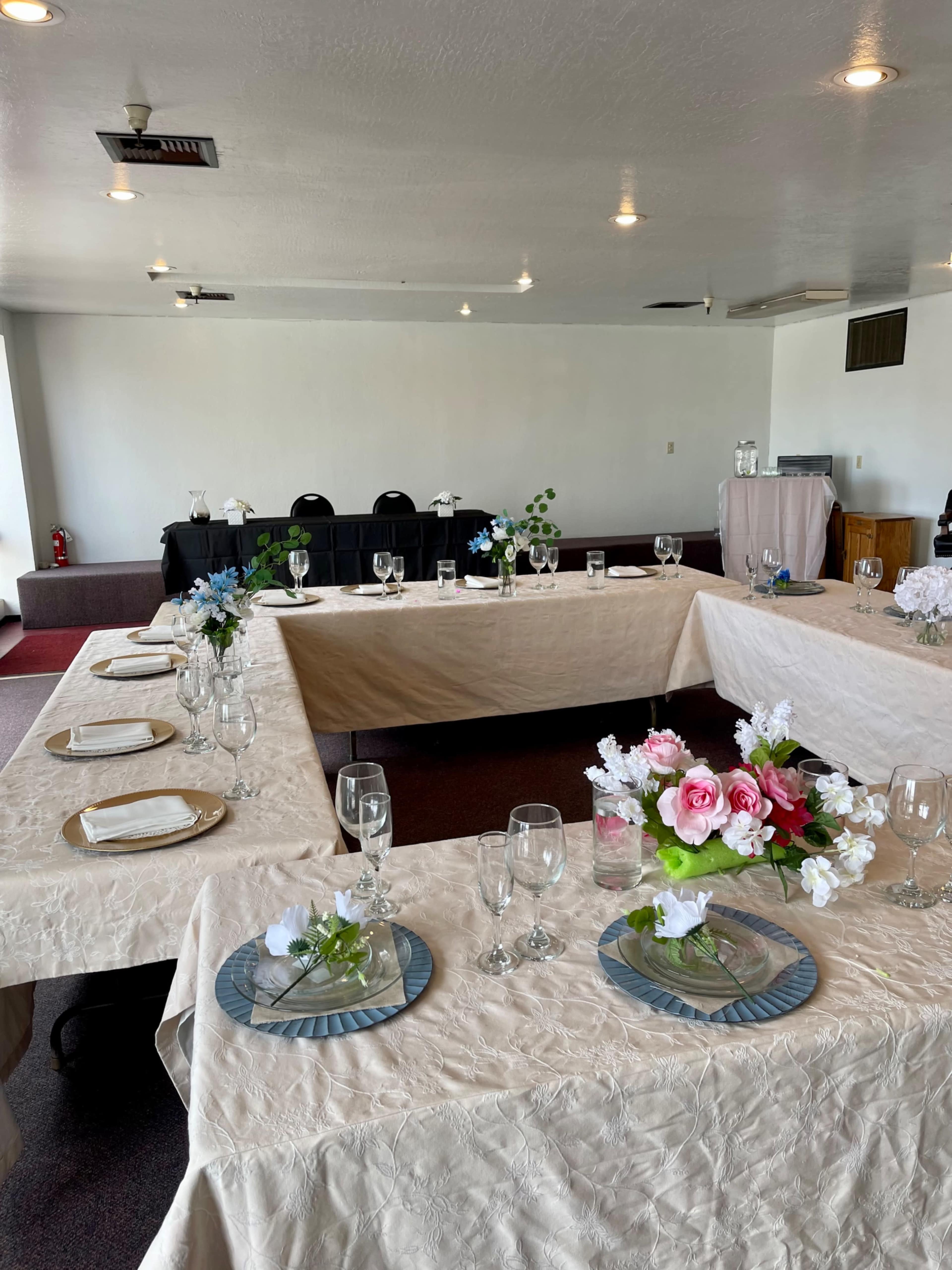 A spacious room with multiple tables arranged in an L-shape, each set with plates, glasses, and floral centerpieces.