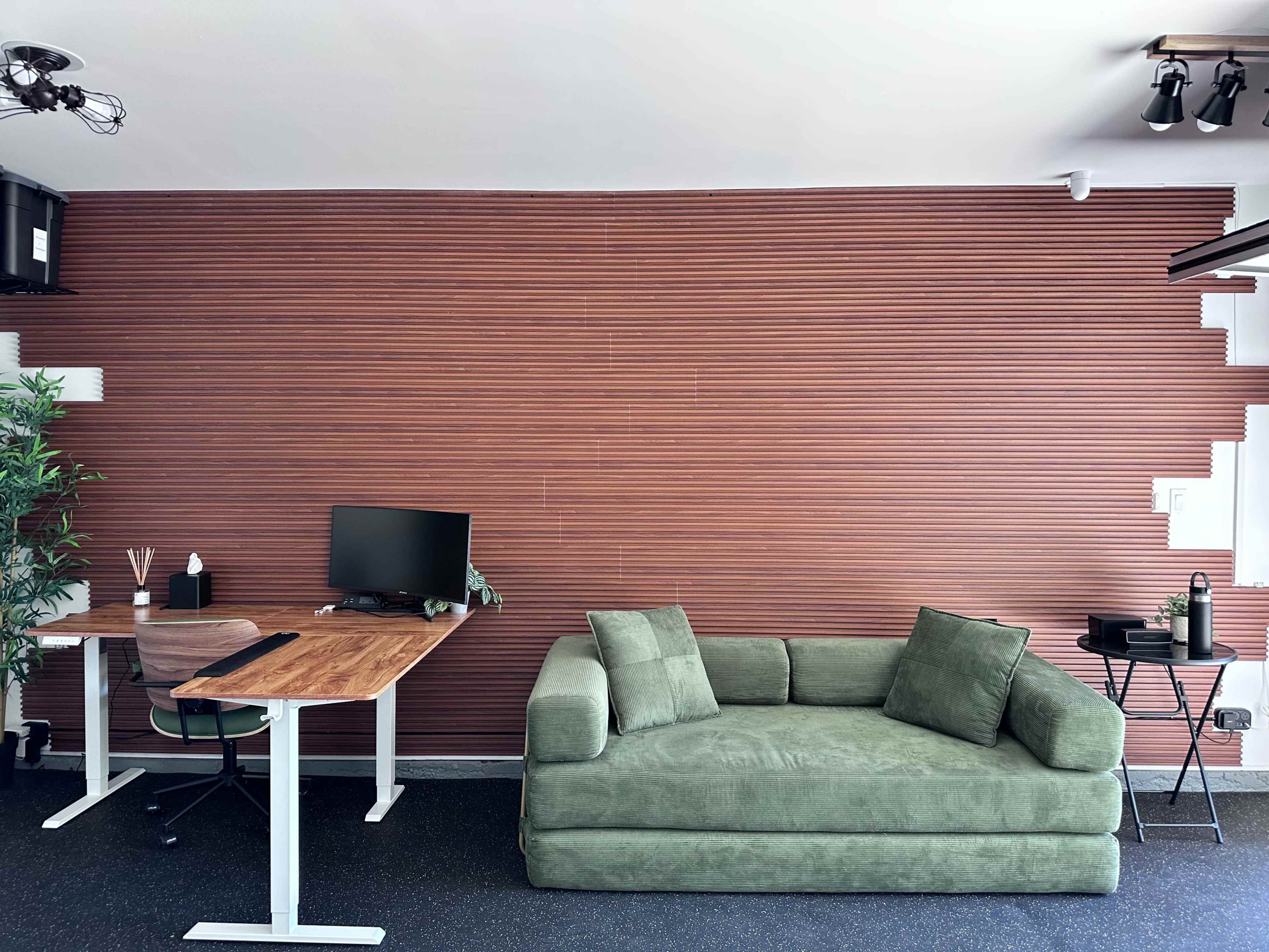 The image shows a modern office space with a green sofa, a wooden desk with a computer monitor, and a potted plant against a textured brown wall.