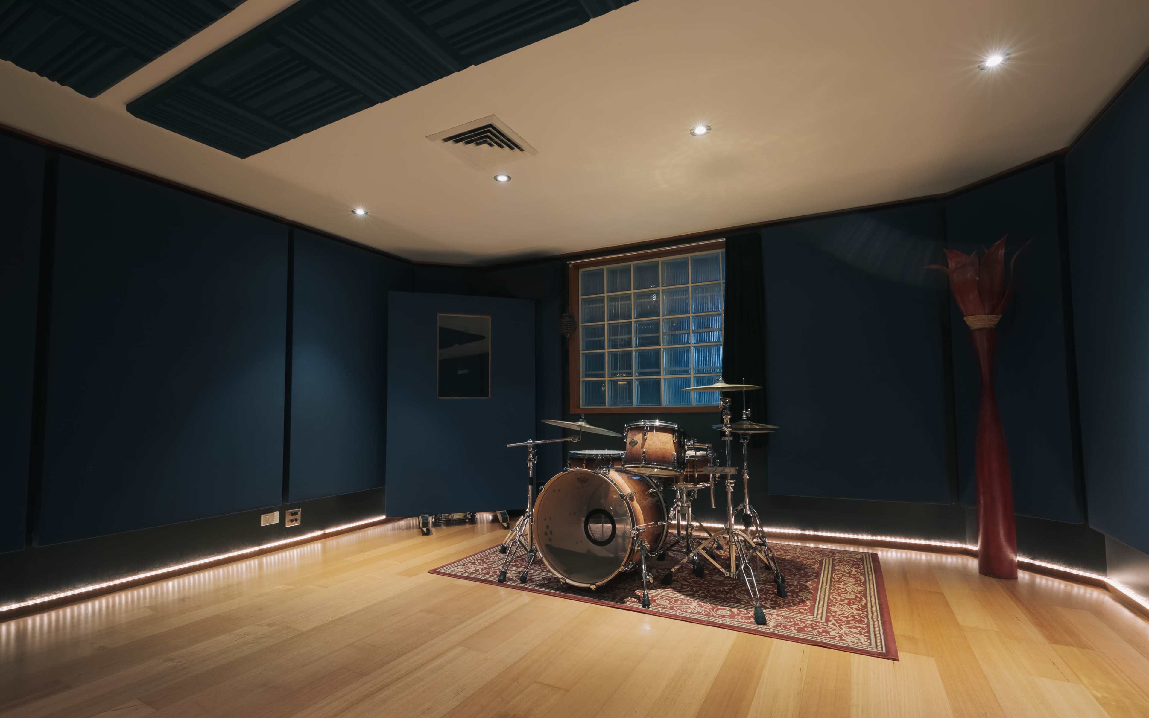 A music recording studio featuring a drum set positioned on a patterned rug in a room with dark blue walls and hardwood flooring.