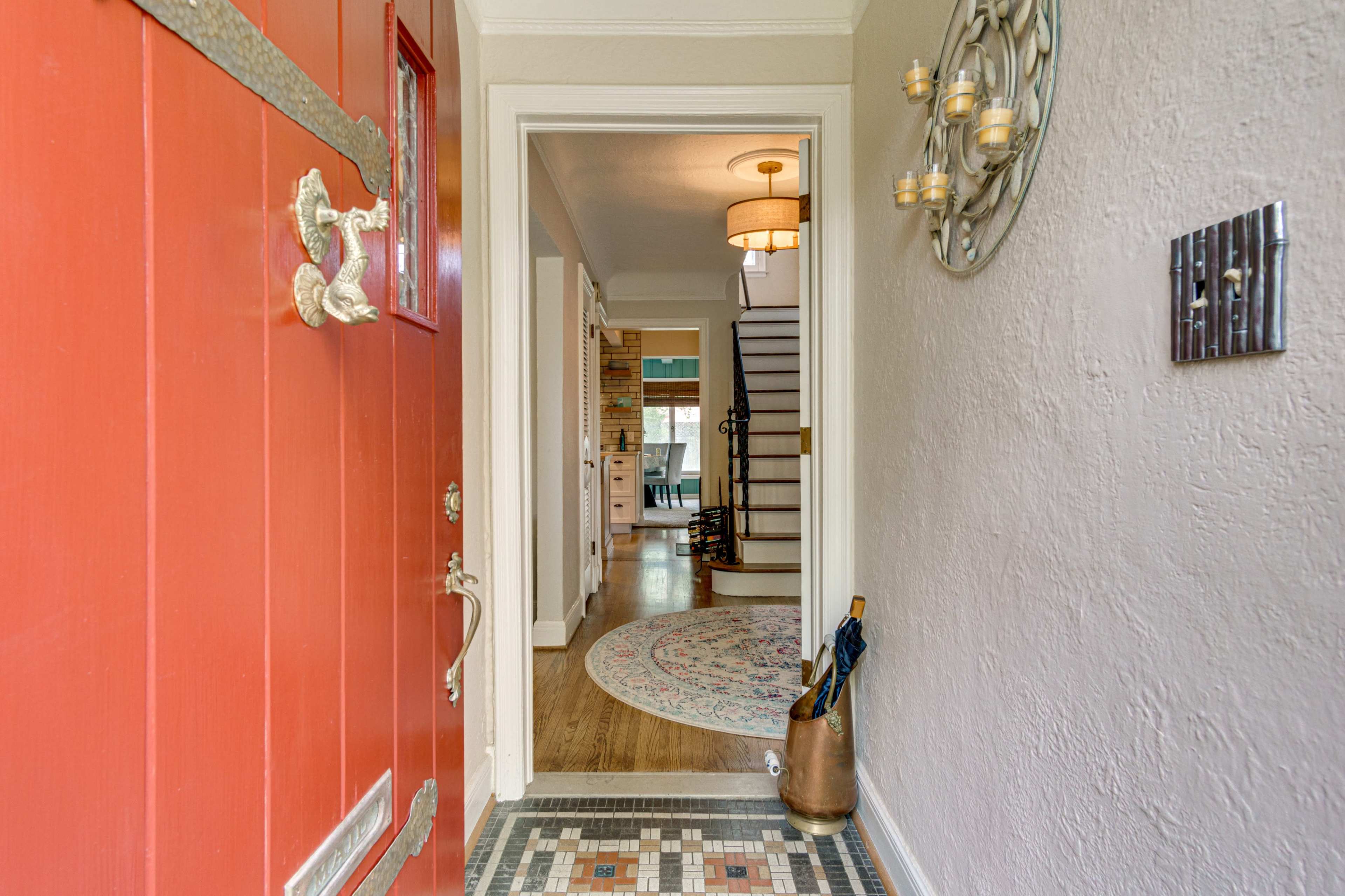 The image shows an inviting entryway featuring a red front door, a patterned tile mat, and a view of a staircase leading to a brightly lit hallway.