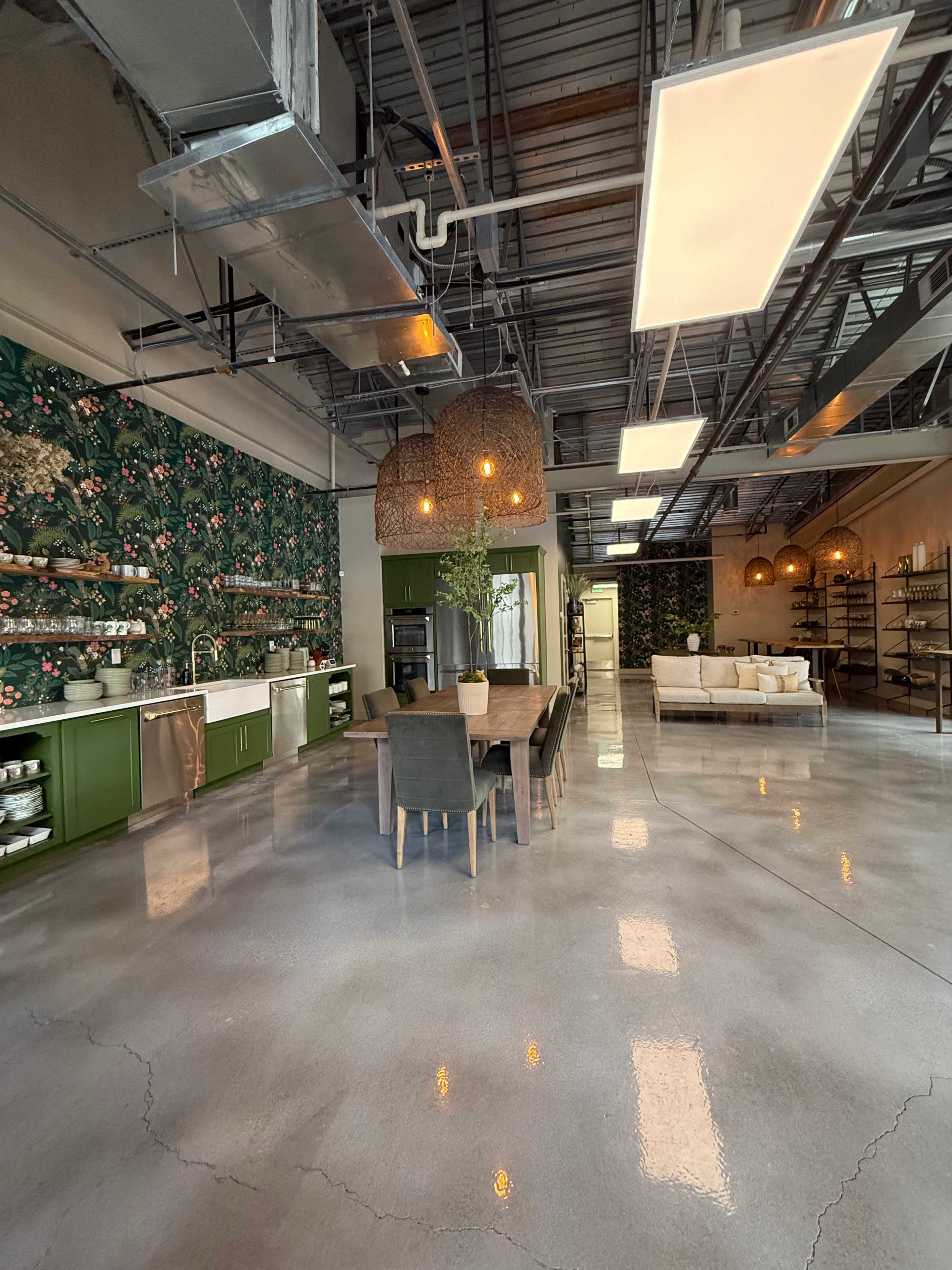 The image shows a spacious kitchen and dining area with green cabinetry, a large wooden dining table, and a living space featuring a light-colored sofa, accented by decorative lighting and floral wallpaper.