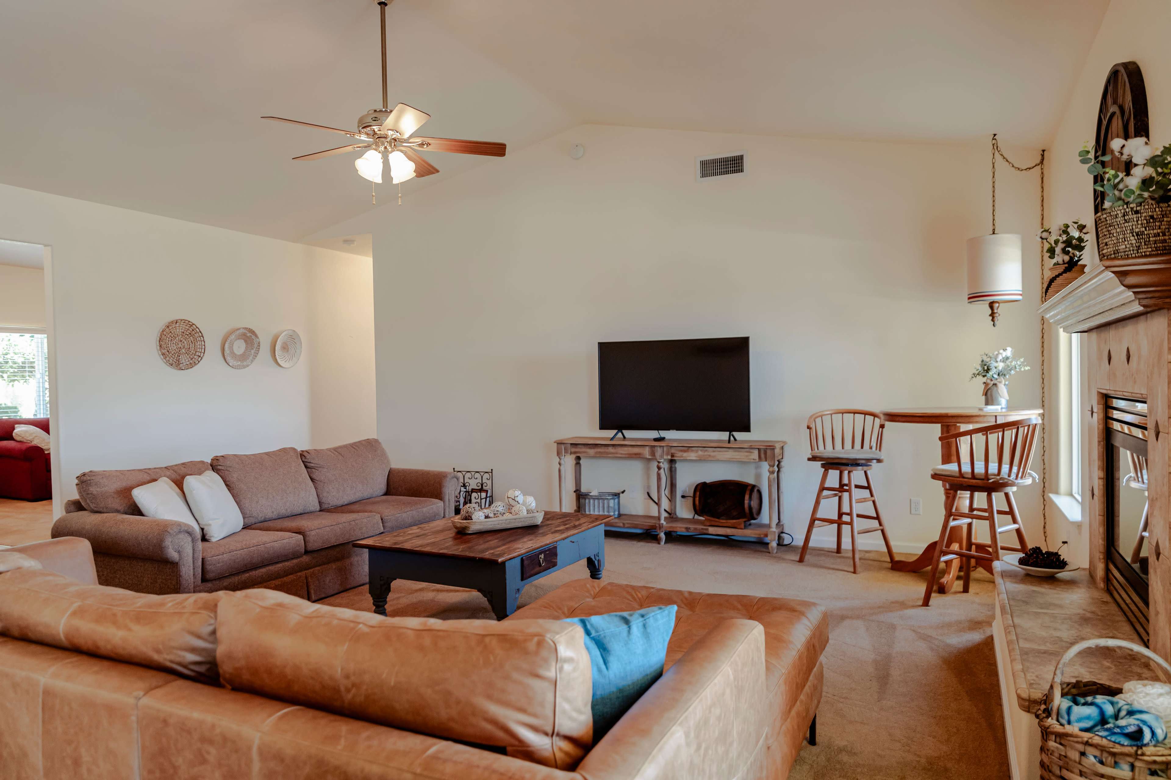 A brightly lit living room featuring two sofas, a coffee table, a television on a stand, and a small dining area with bar stools.