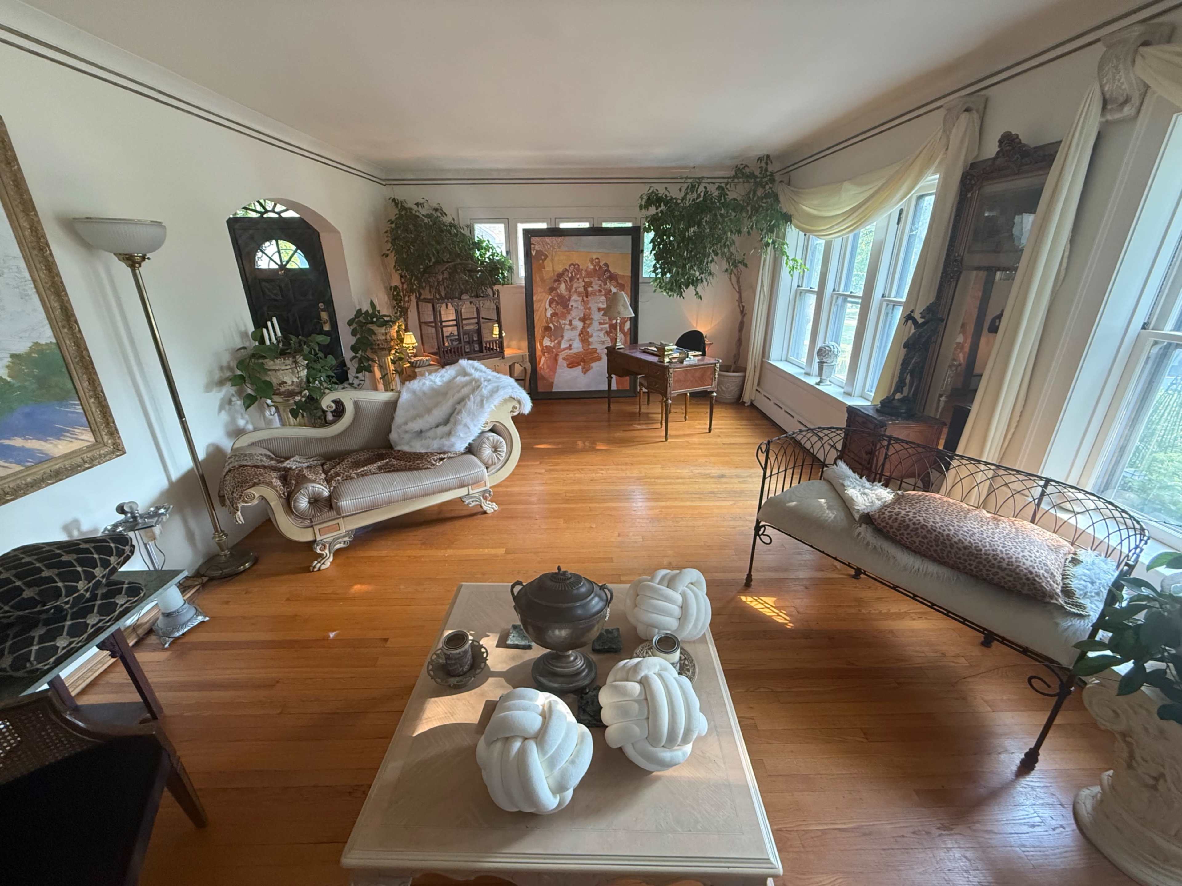 The image shows a spacious living room furnished with a vintage couch, a coffee table with decorative items, and large windows allowing natural light to fill the space.