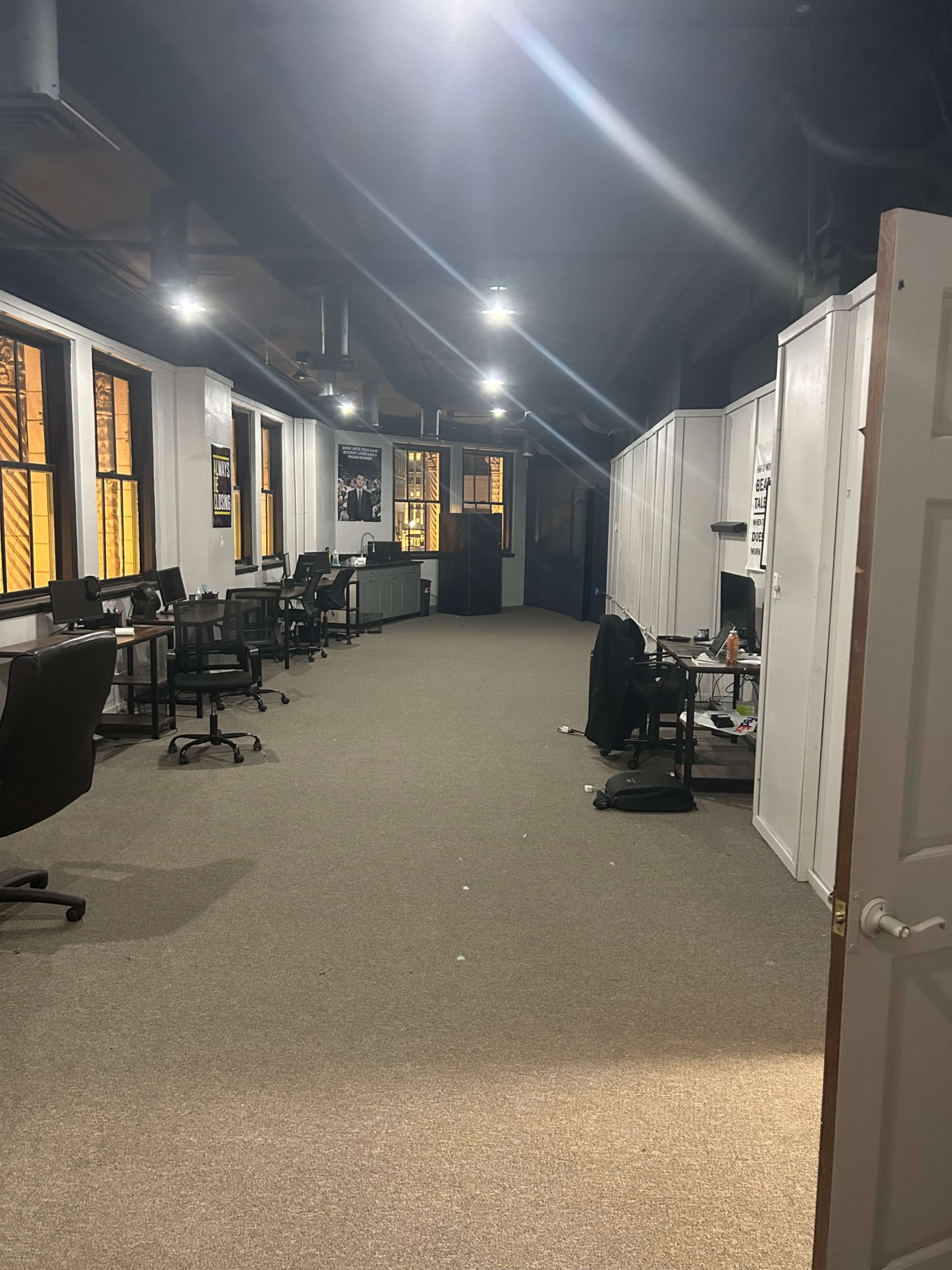 A dimly lit office corridor with several chairs and desks on either side, leading to a darker area at the far end.