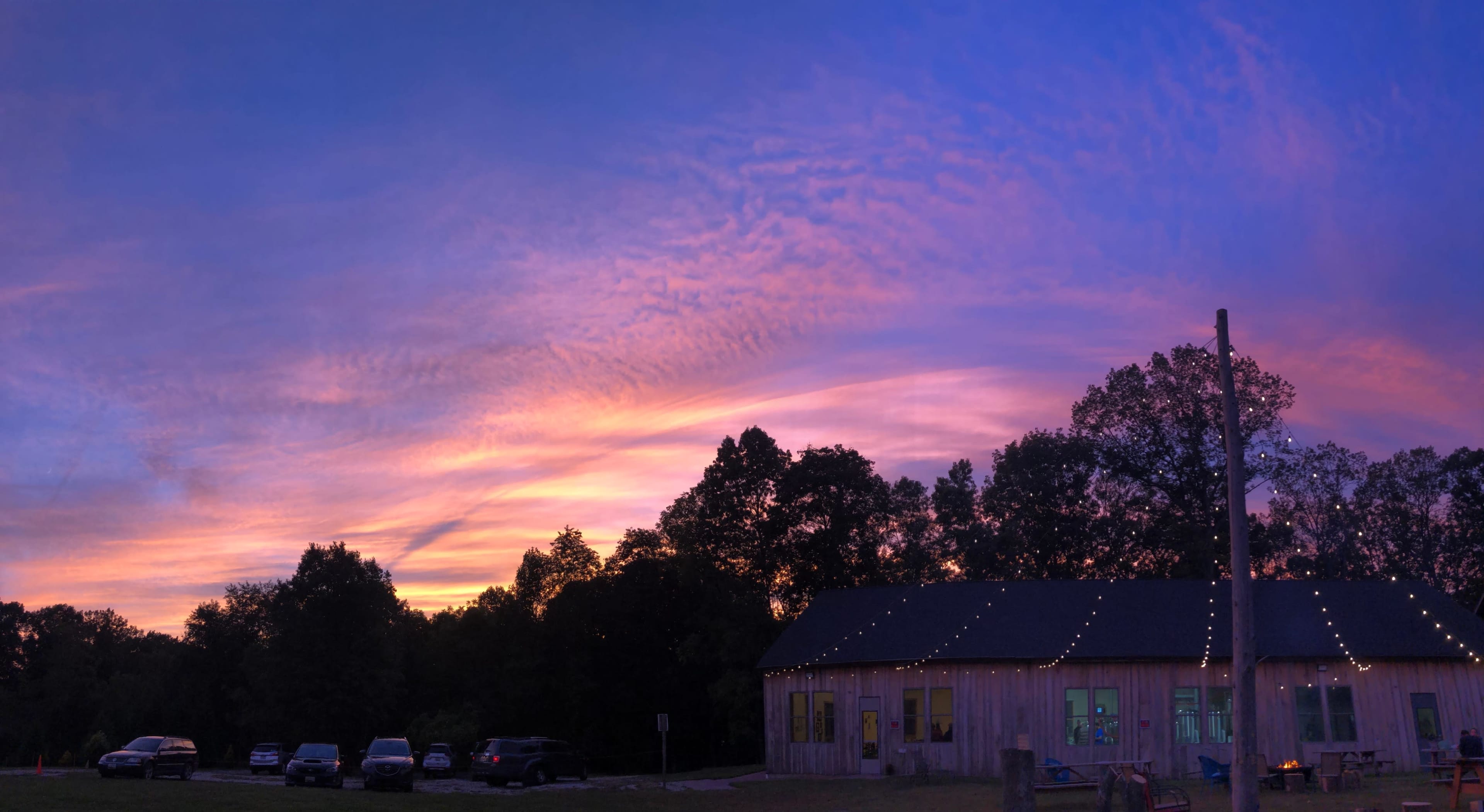A wooden building with string lights sits under a colorful sunset, surrounded by trees and parked cars.