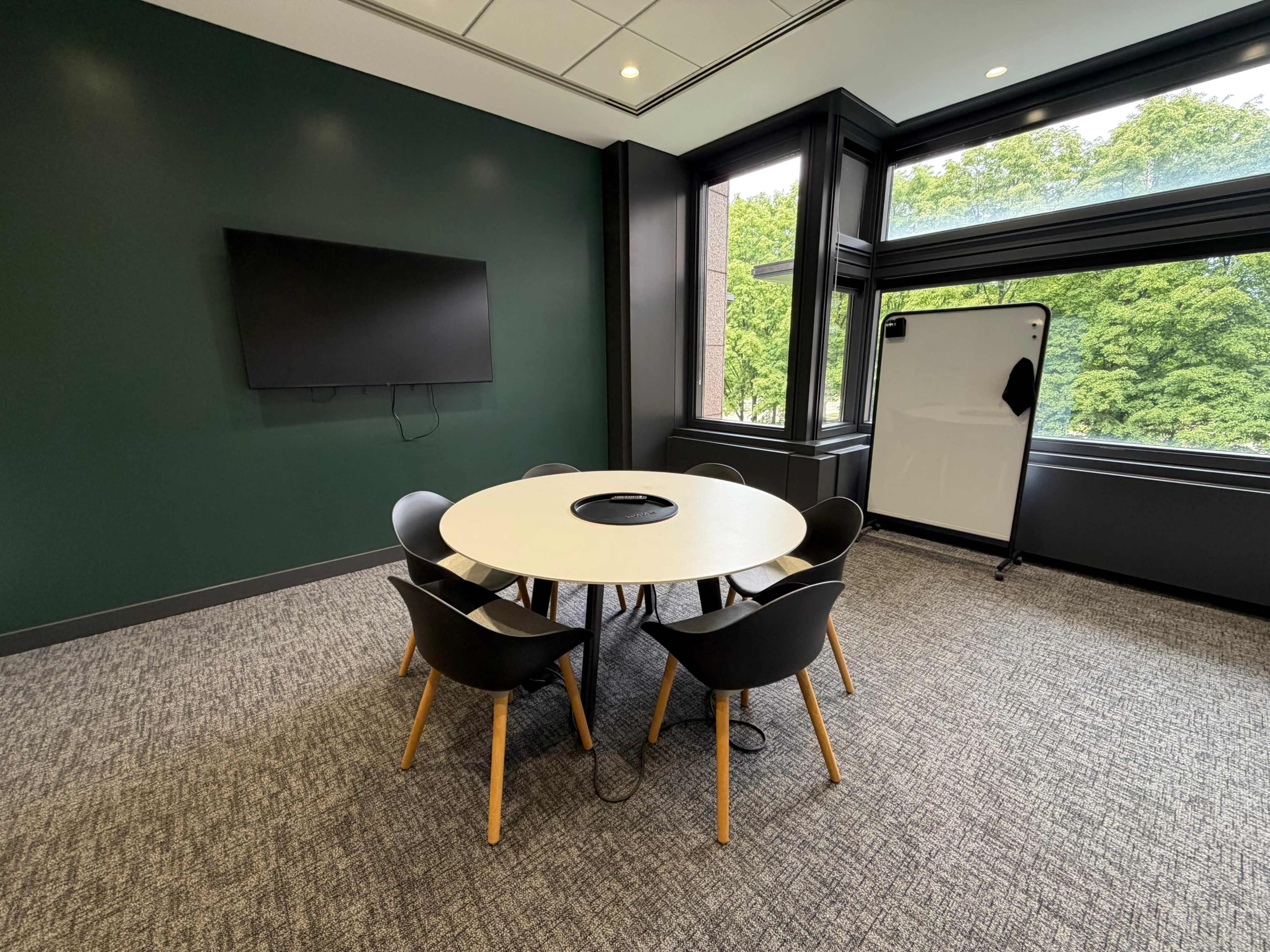 A conference room features a circular table surrounded by five chairs, a wall-mounted screen, and a movable whiteboard next to a large window with views of greenery outside.