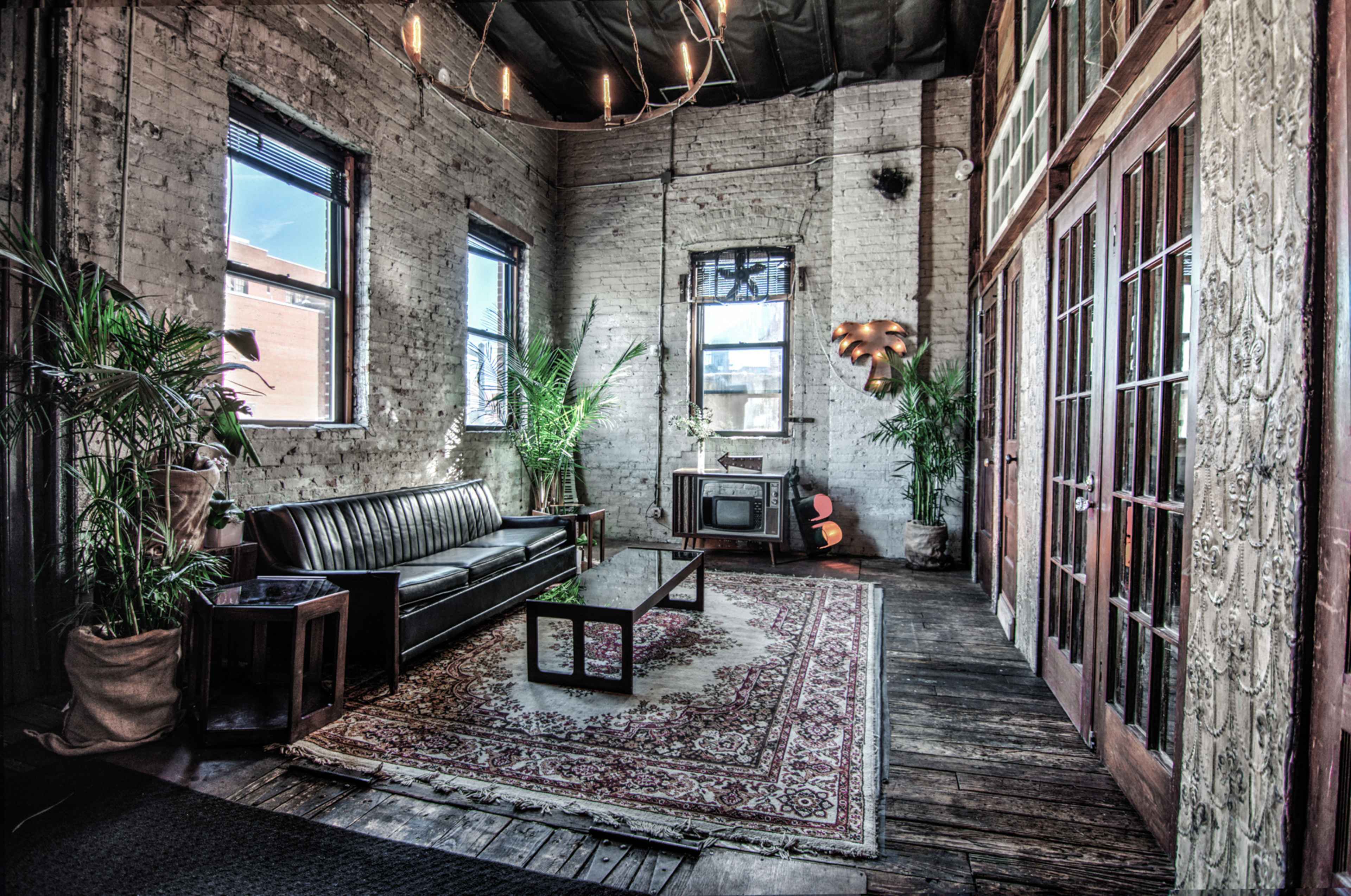 A spacious room with exposed brick walls, a leather sofa, a coffee table on a patterned rug, and large windows allowing natural light to enter.