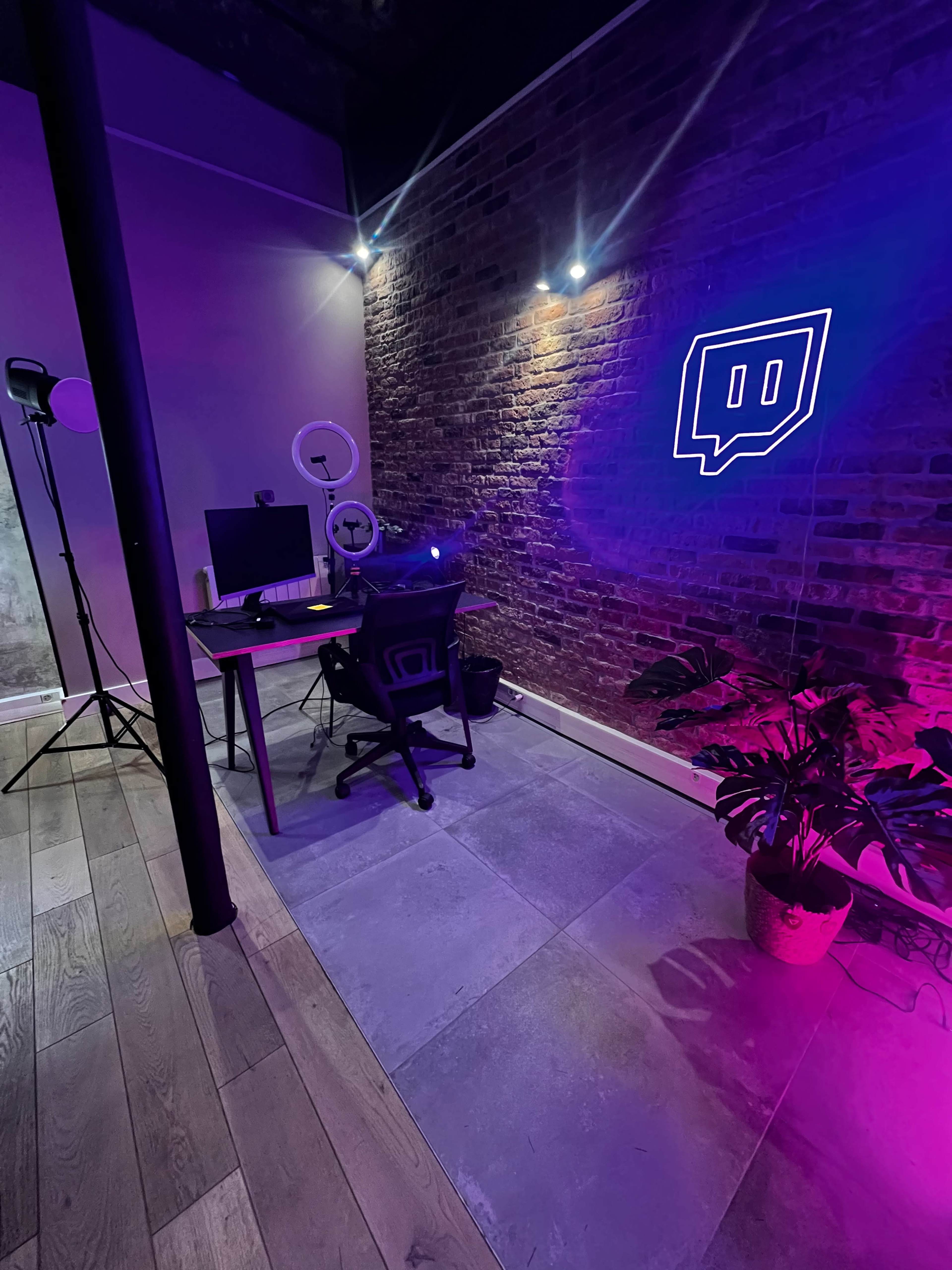 The image shows a modern workspace featuring a desk with a computer, illuminated by purple lights and a neon Twitch logo against a brick wall.