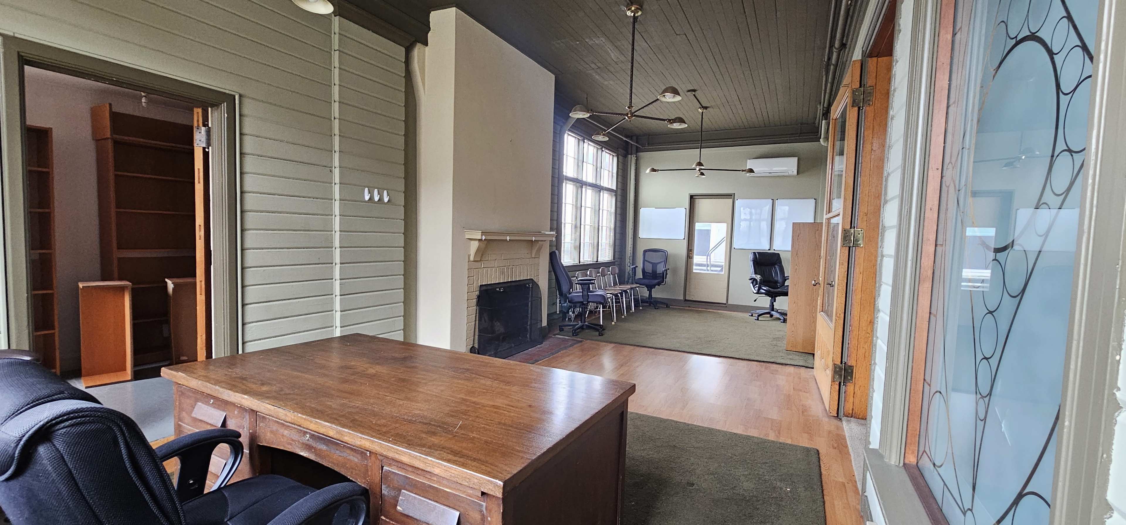 An office space with wooden flooring, a large desk, and several chairs arranged around a fireplace and windows.