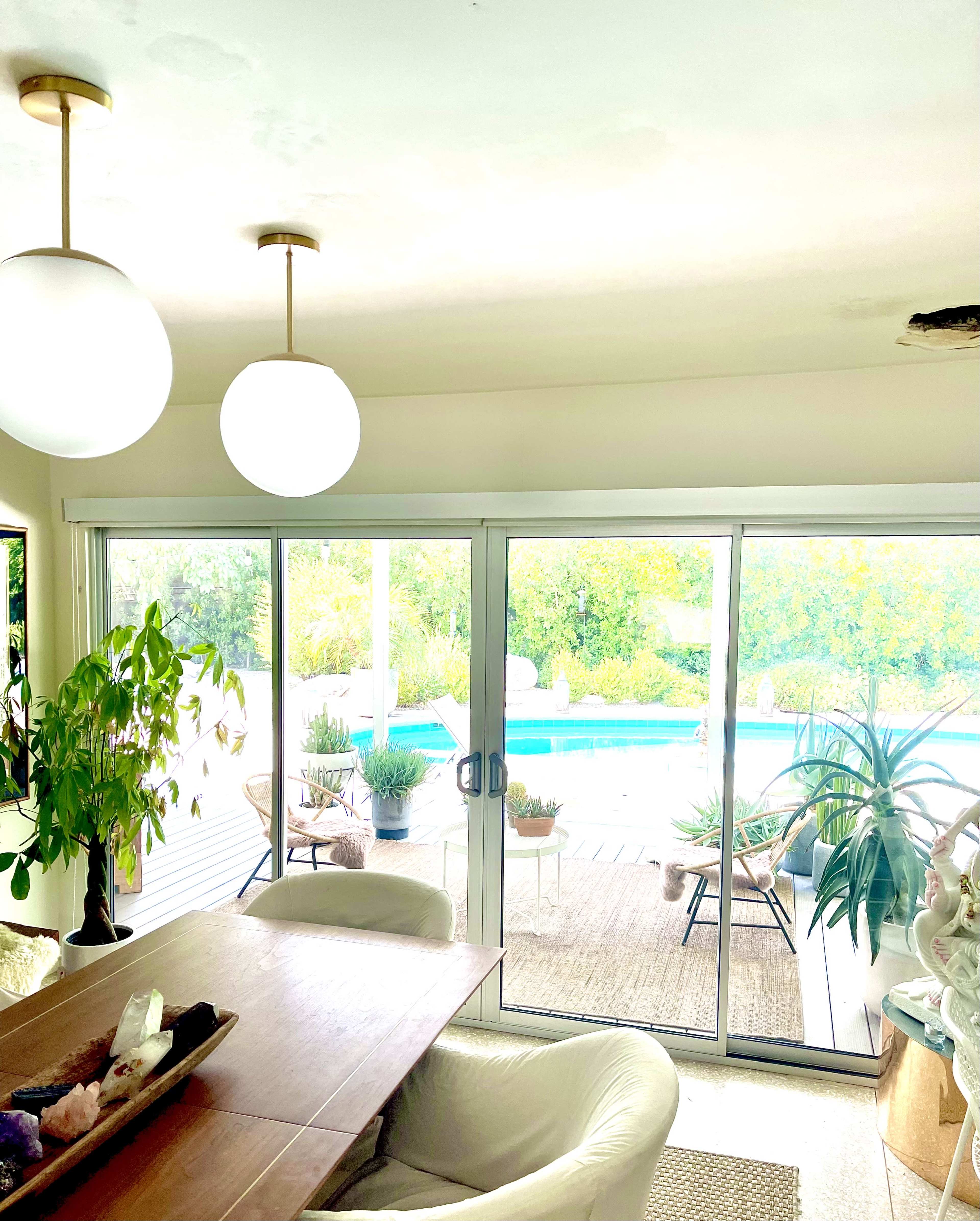 The image shows a dining area with a wooden table and two hanging light fixtures, leading to sliding glass doors that open to a pool and patio area.