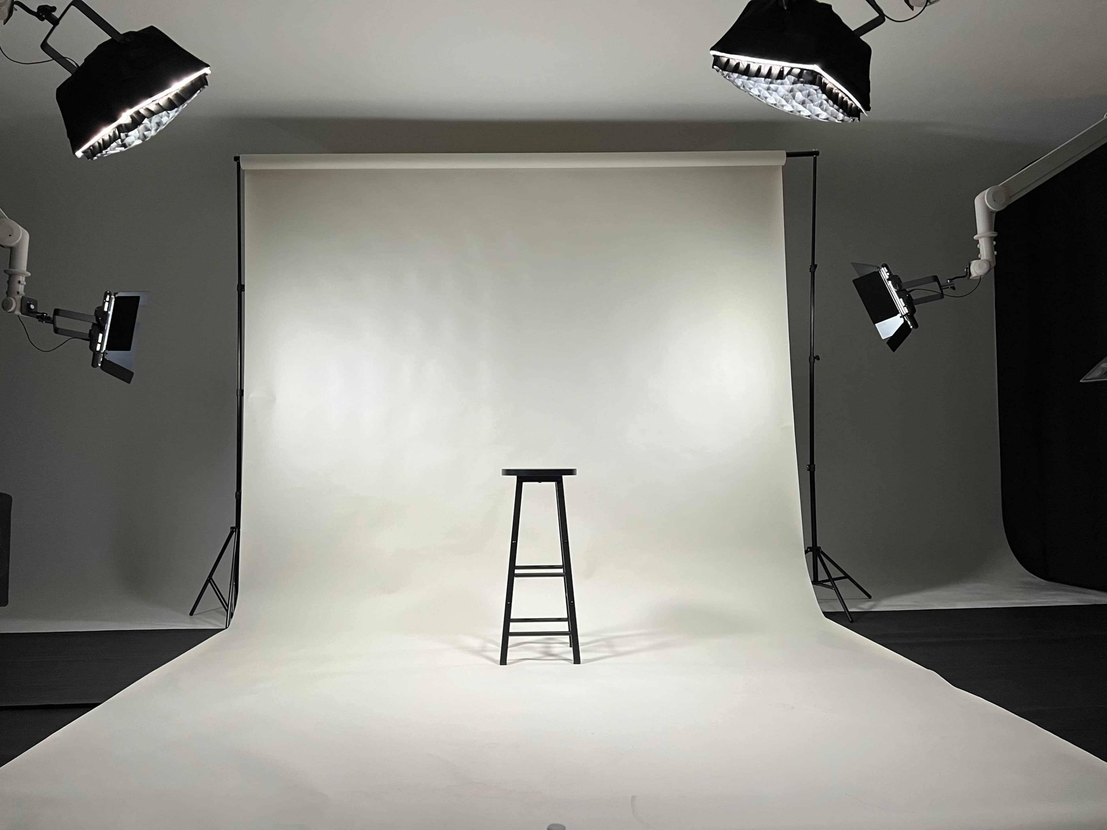 A photography studio setup with a simple black stool positioned on a seamless white backdrop, illuminated by overhead lights.