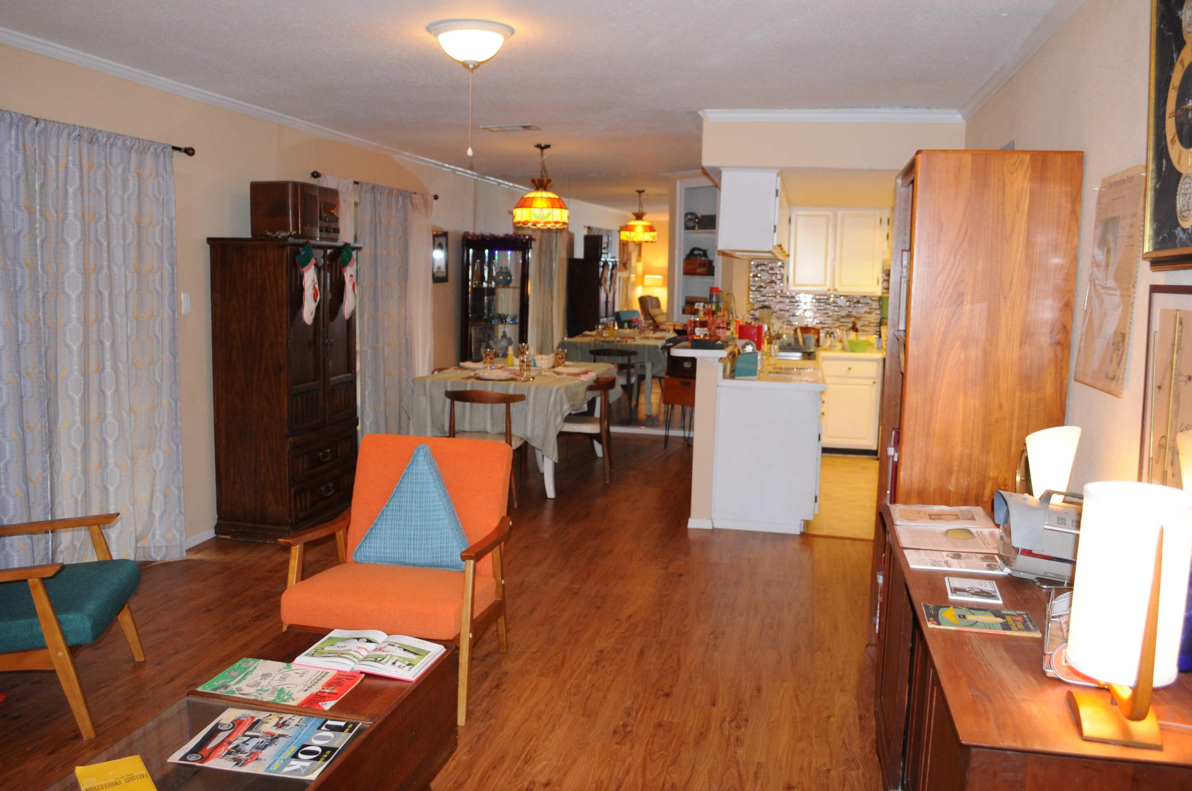 The image shows a living space with a seating area, a dining table, and an open kitchen, featuring wooden furniture and warm lighting.
