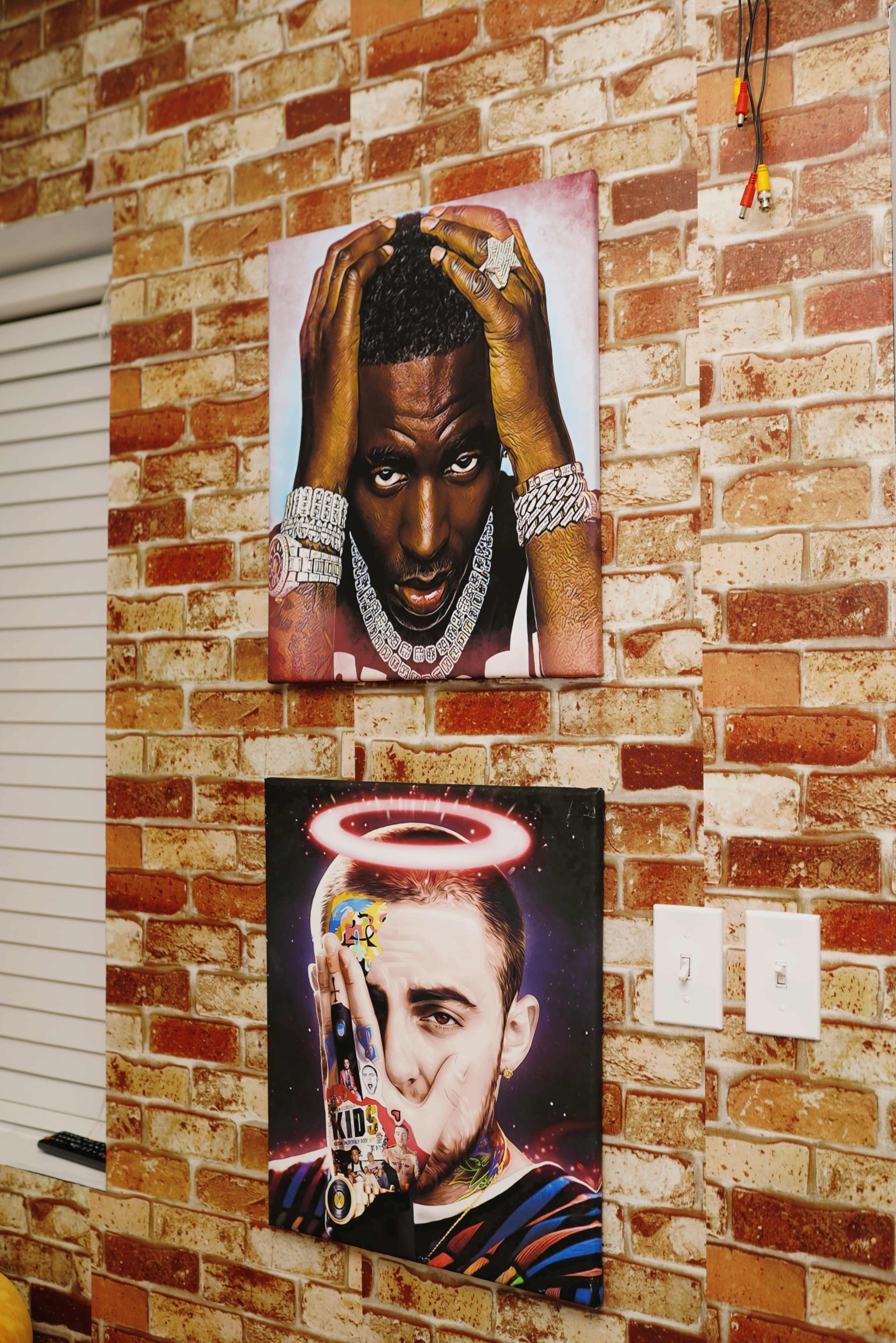 Two framed portraits hang on a brick wall; one features a man with jewelry and a serious expression, while the other depicts a man with an illuminated halo and a thoughtful gesture.