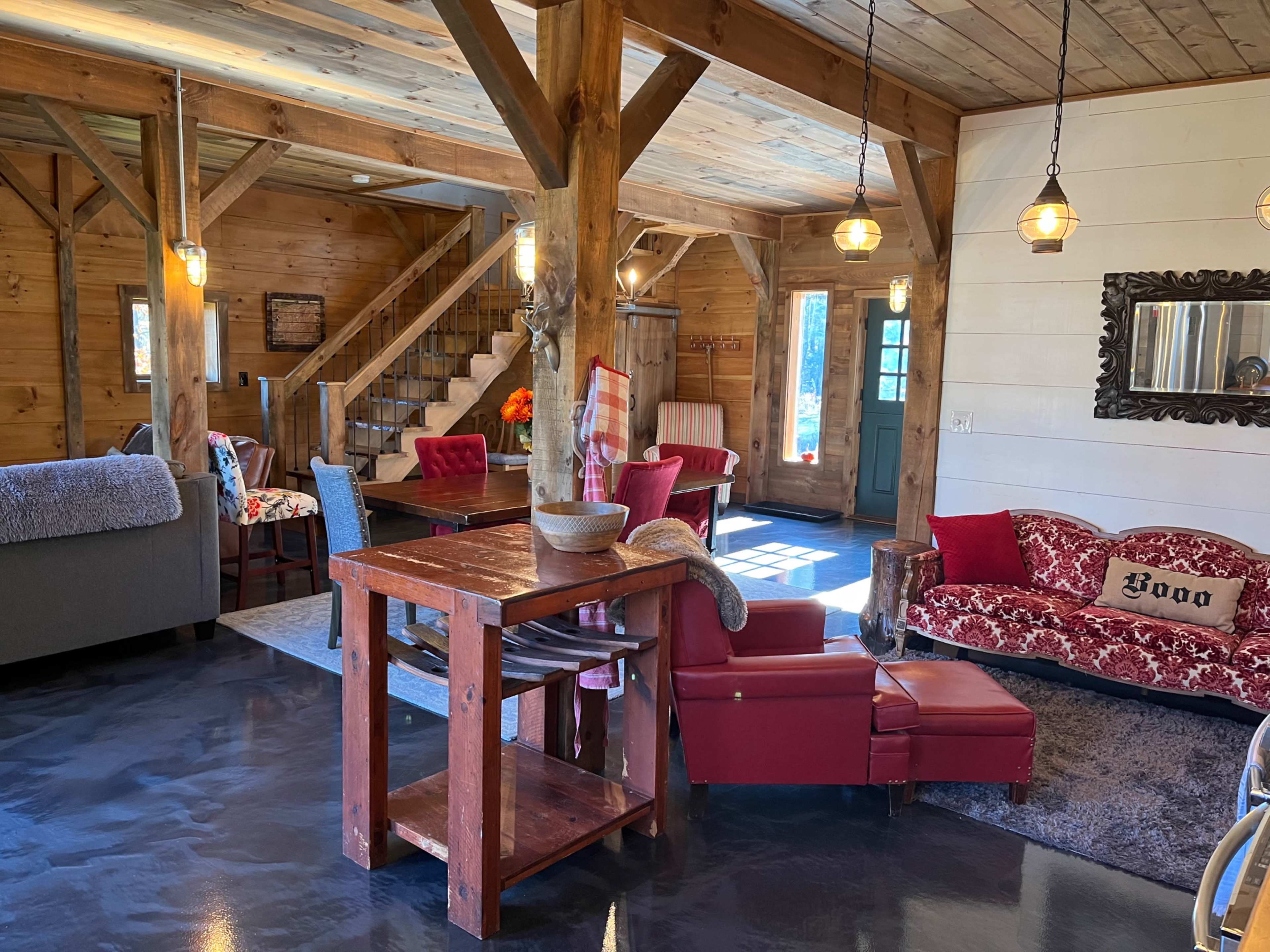 A bright, rustic interior of a wooden cabin featuring a mix of red and cream furniture, a dining area, and a staircase leading to an upper level.