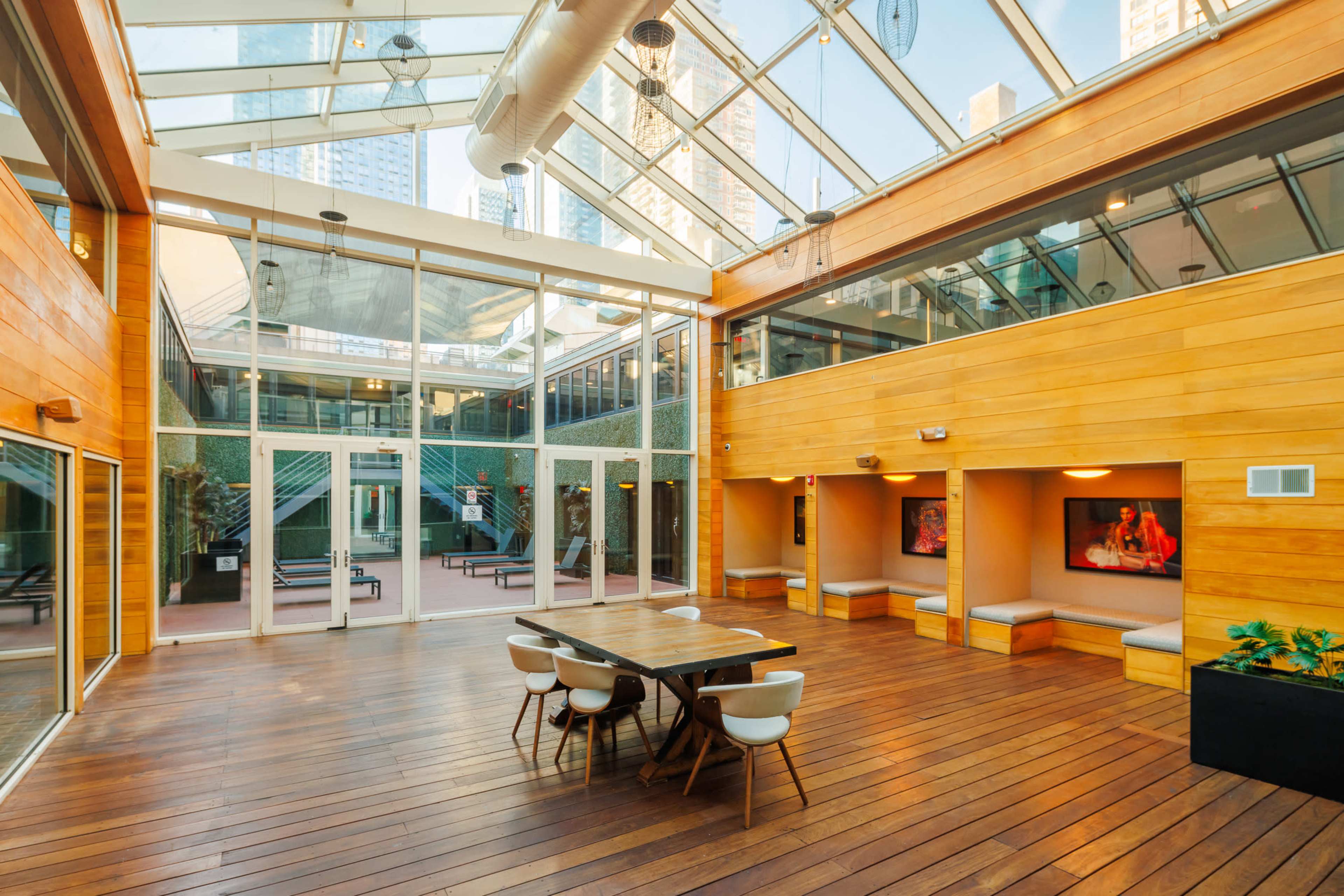 The image shows a spacious, brightly lit indoor area with wooden flooring, large glass windows, and a seating arrangement along the walls.
