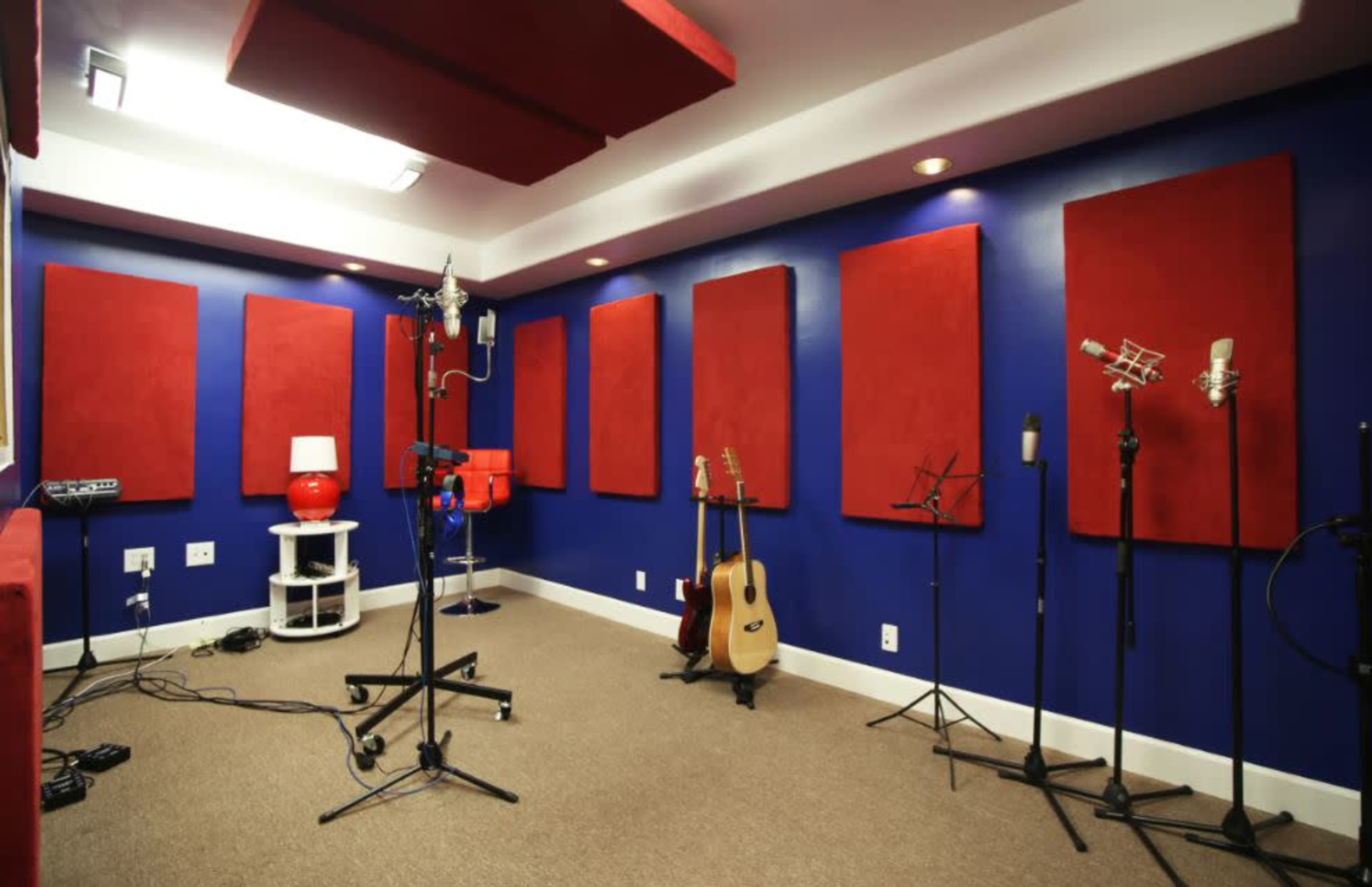 The image shows a sound studio featuring blue walls, red acoustic panels, various microphones on stands, a guitar, and a lamp on a white table.