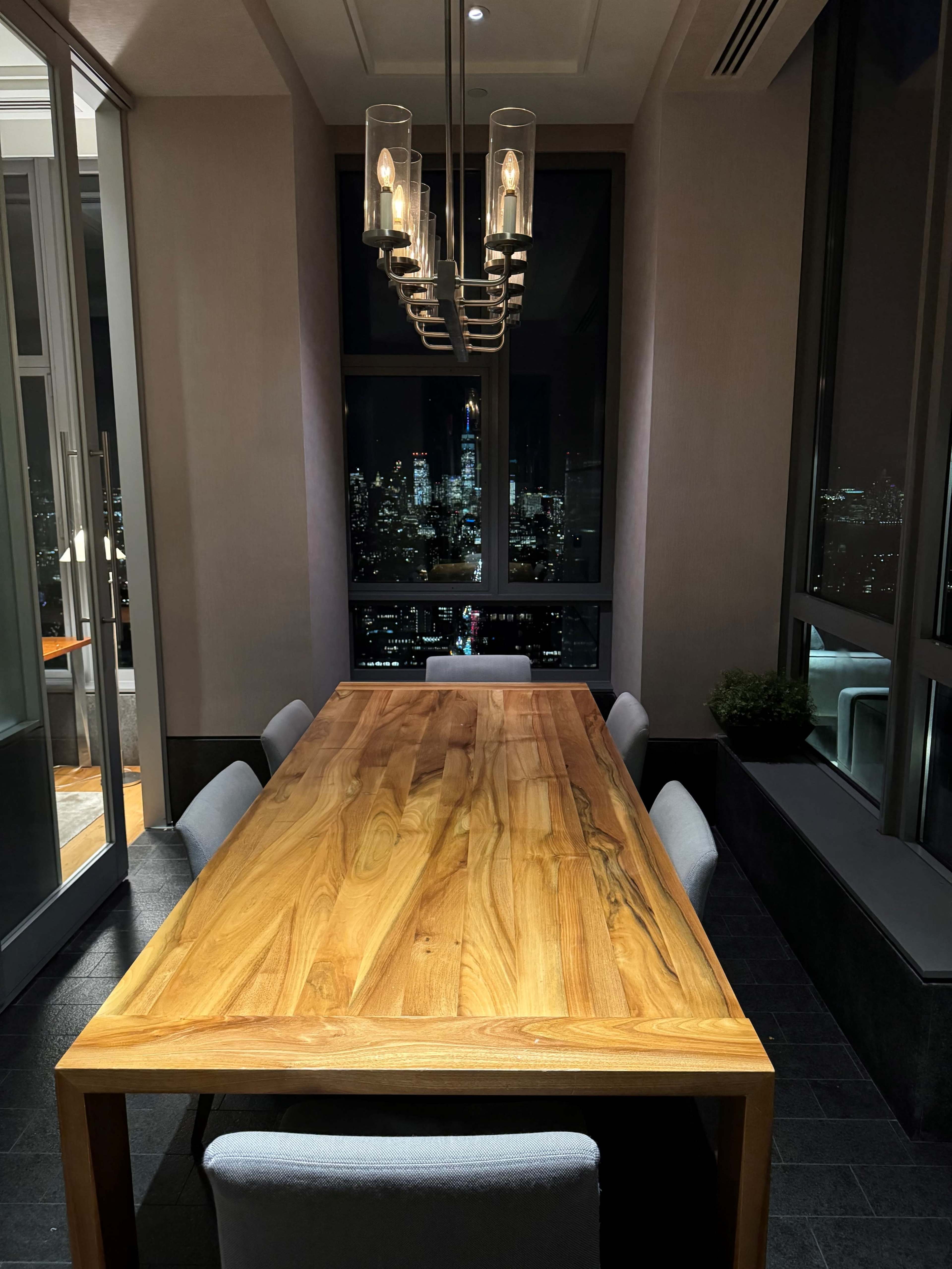A wooden dining table with six chairs sits in a dimly lit room featuring large windows that showcase a city skyline at night.