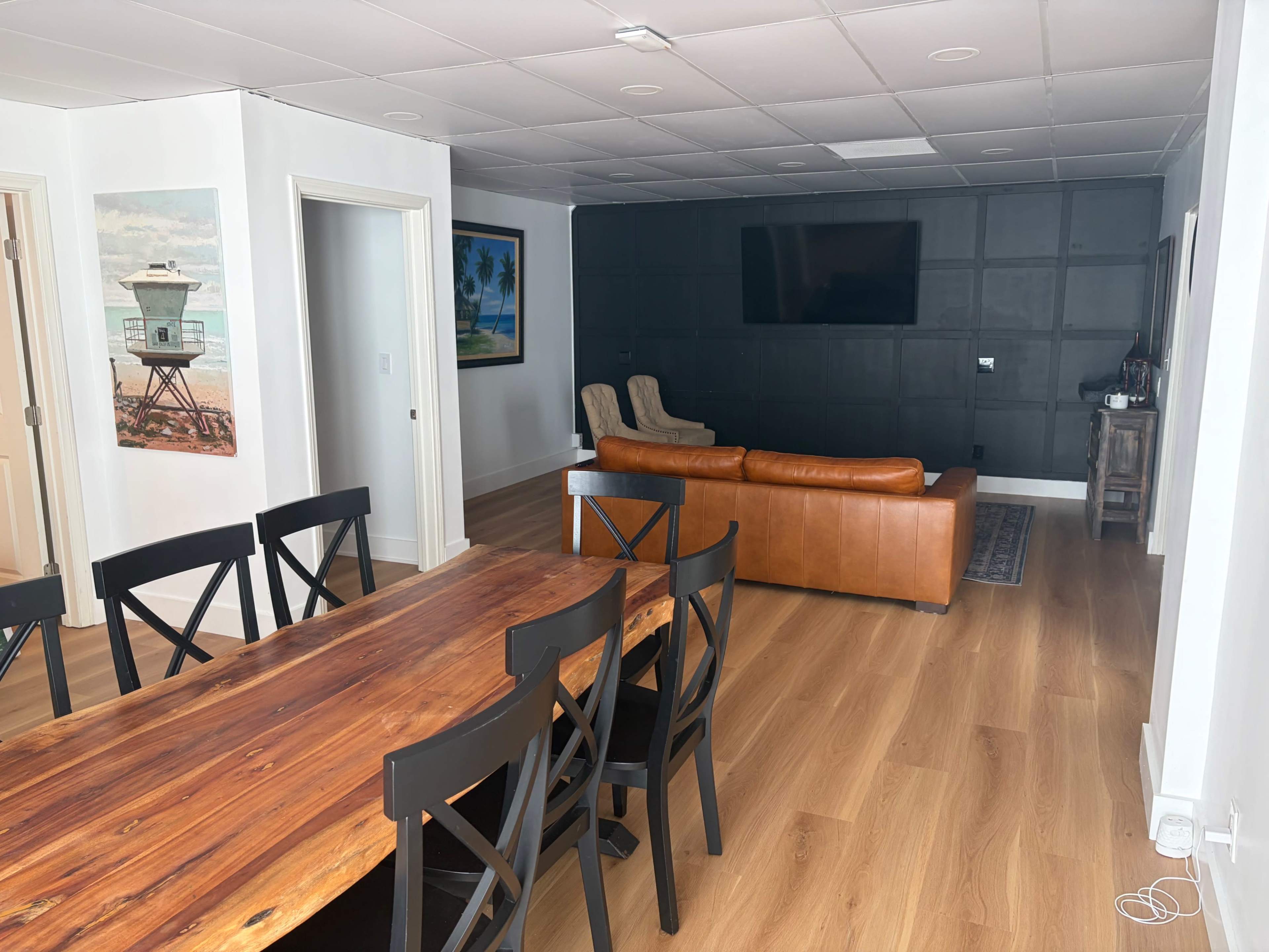 The image shows a spacious interior with a wooden dining table and black chairs on the left, while a living area featuring a brown leather sofa and a wall-mounted TV is located on the right.