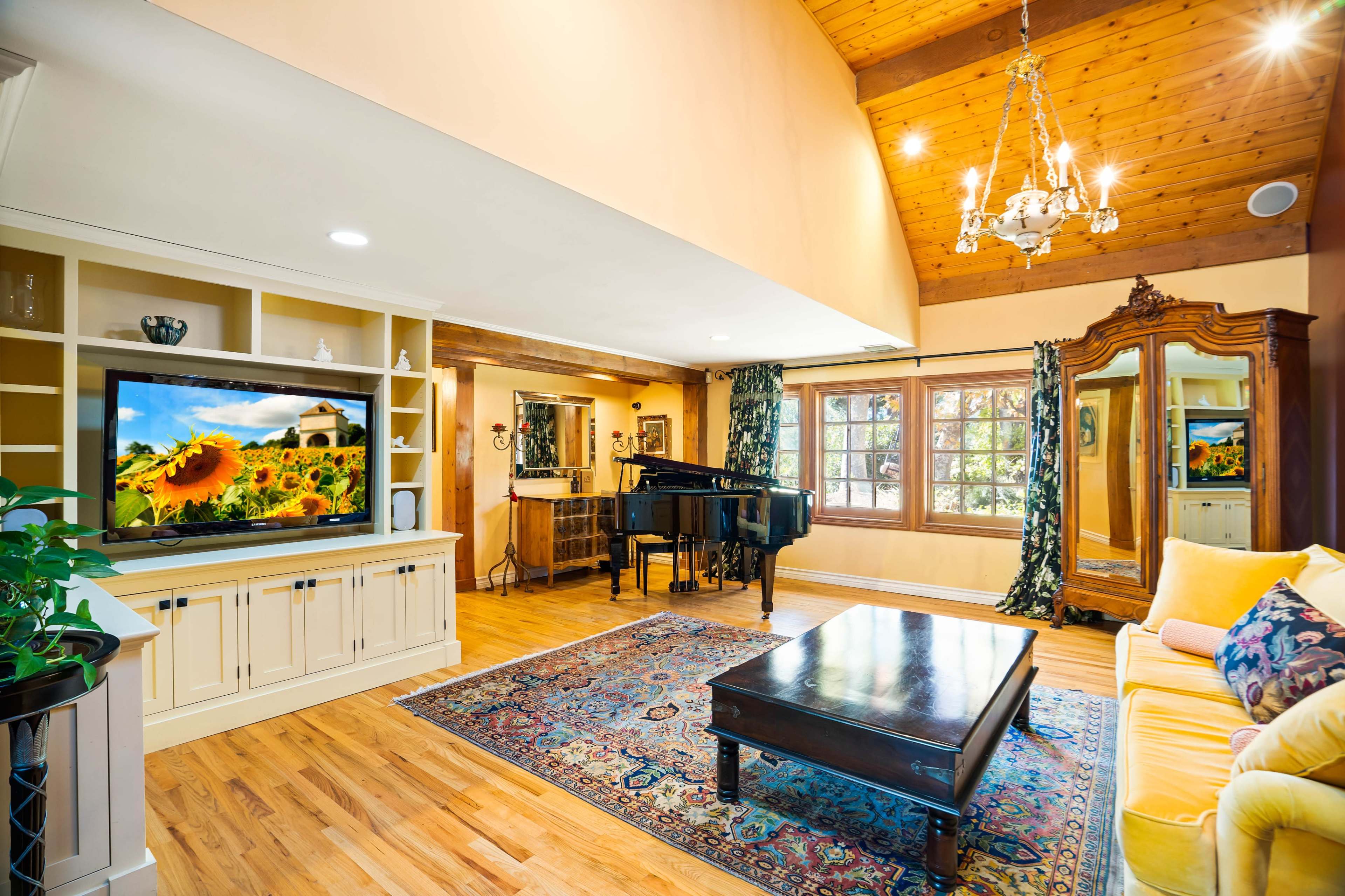 The living room features a grand piano, a large television displaying a scenic image, and elegant furnishings under a high wooden ceiling.