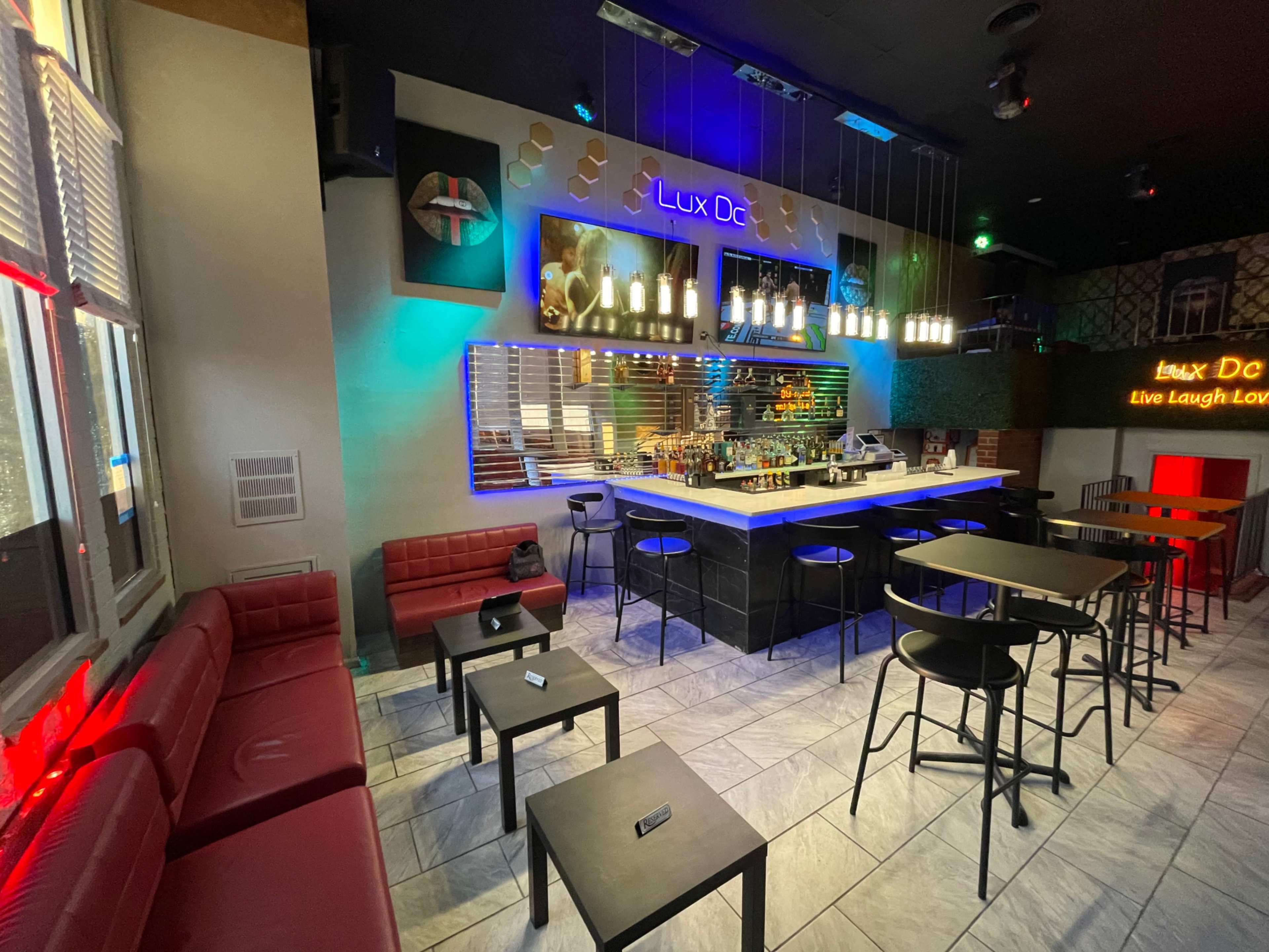 The image shows a modern bar area featuring a sleek counter, seating arrangements with dark tables and red couches, and colorful lighting effects.