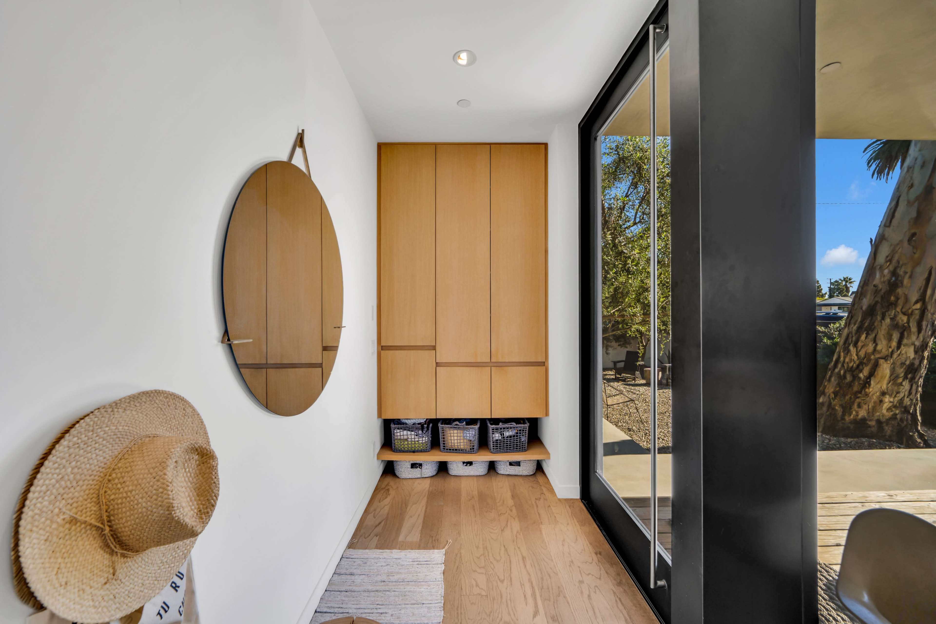 A bright hallway featuring a round mirror, wooden cabinetry, and woven baskets beneath, with a large glass door opening to an outdoor space.