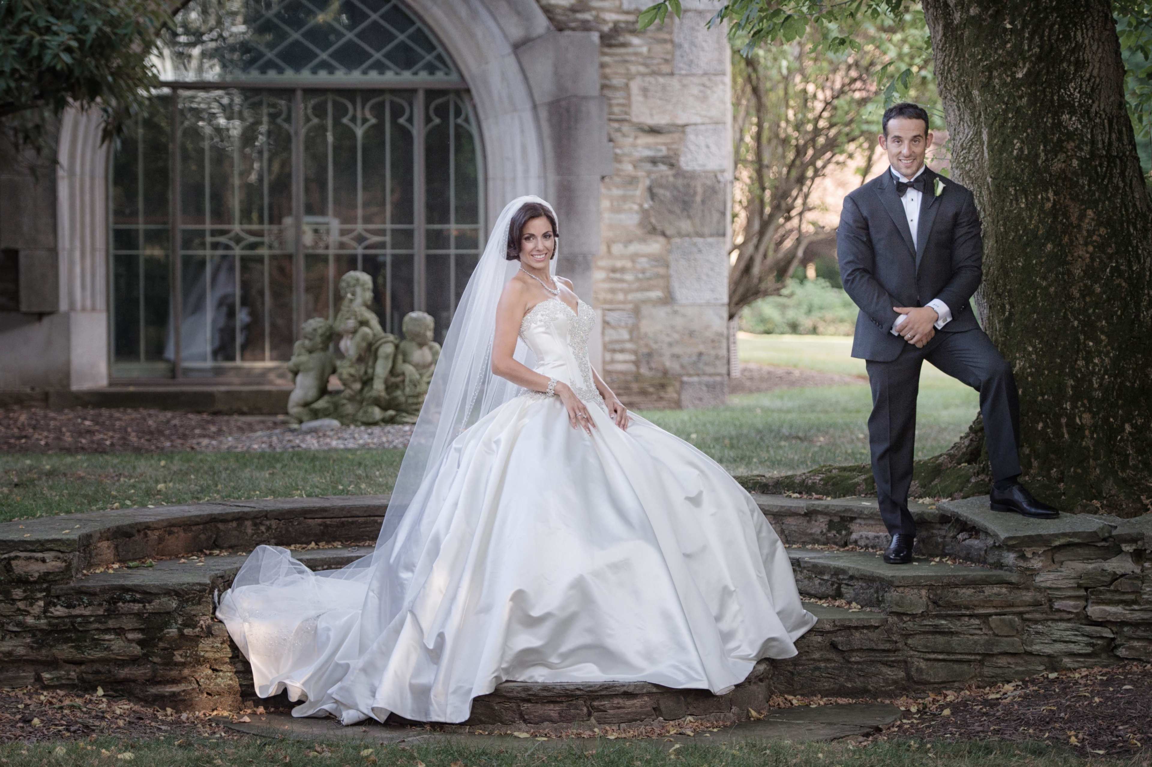 A bride in a white wedding dress and a groom in a black tuxedo pose together in a garden setting near a stone wall and greenery.