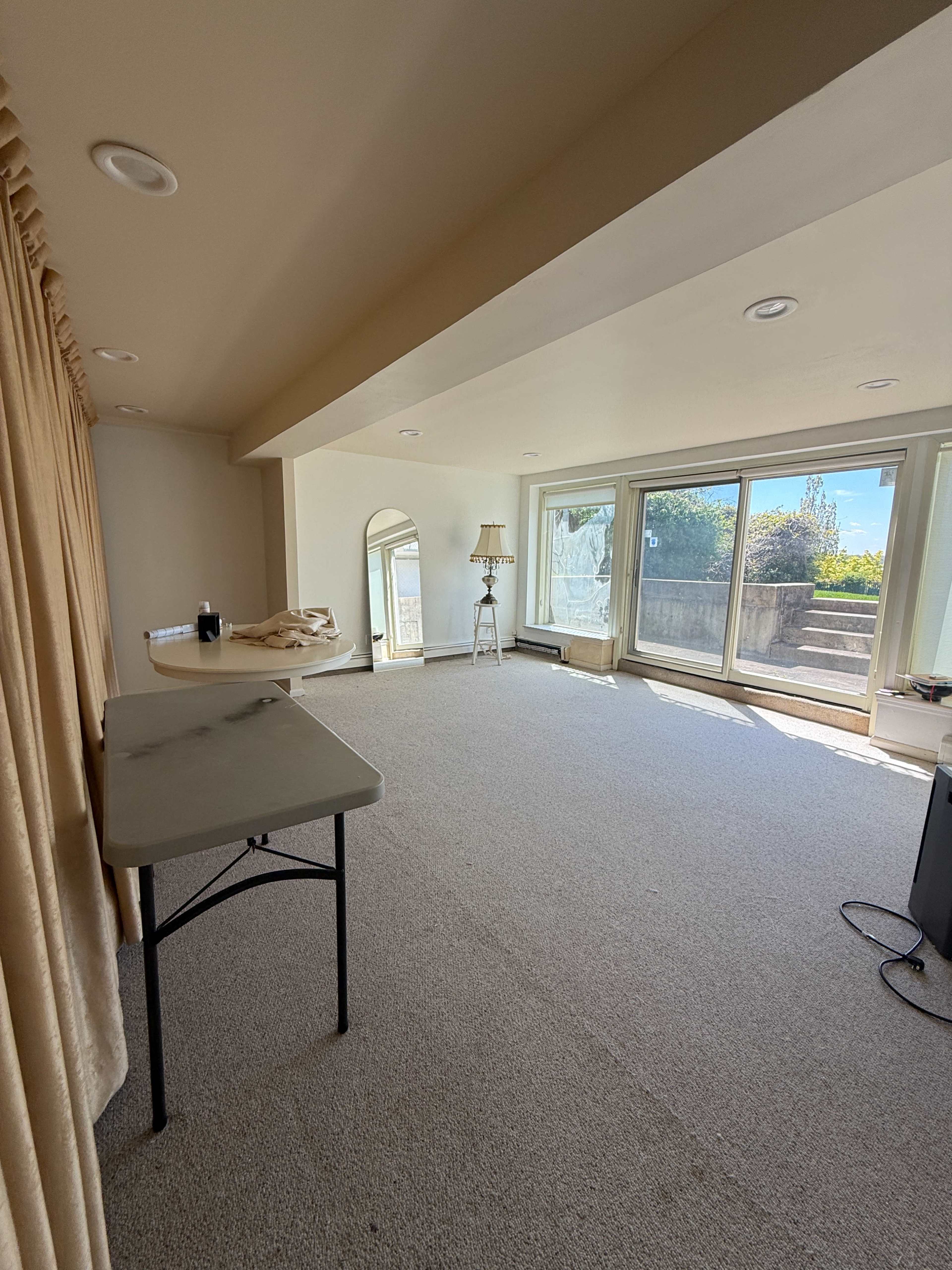 The image shows a spacious, empty room with large windows allowing natural light, a table on the left, and beige carpet covering the floor.
