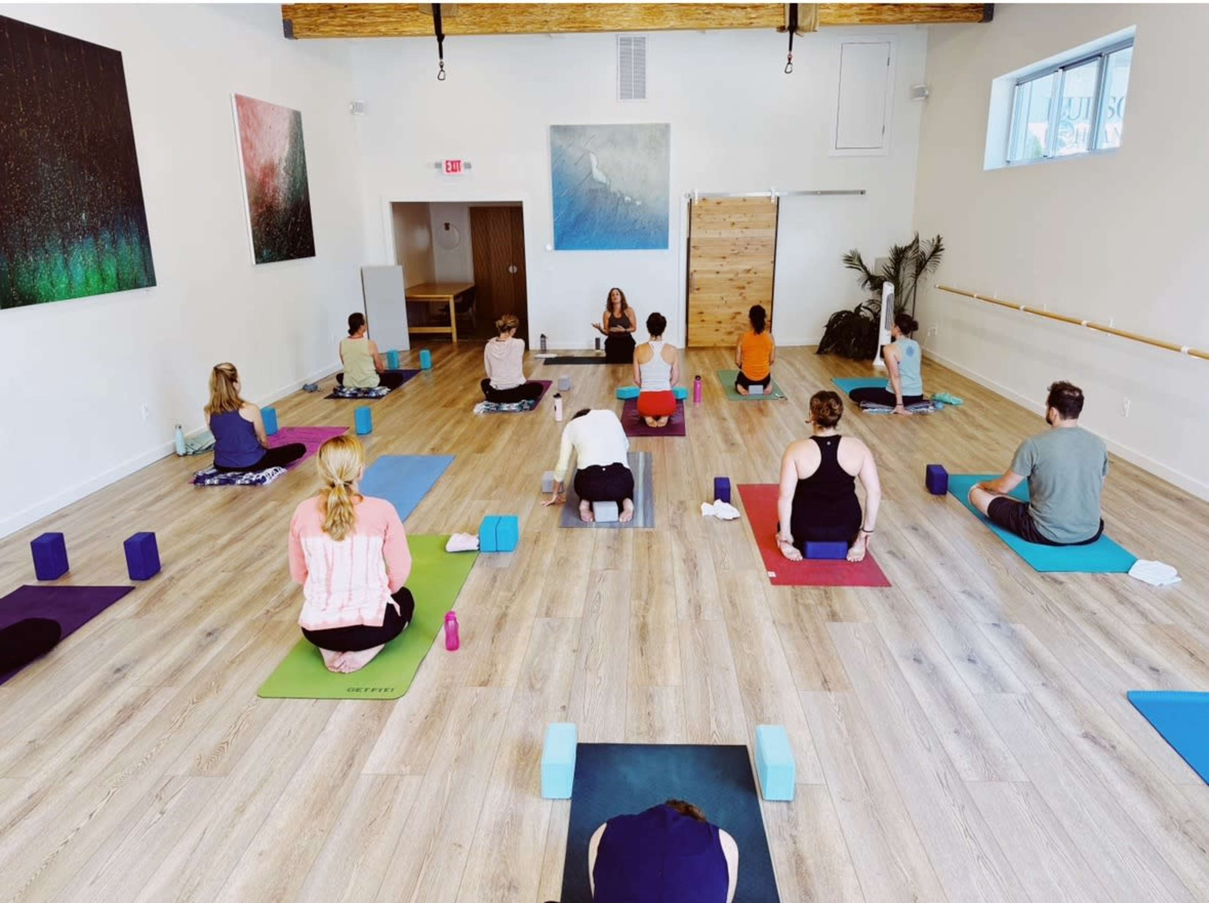 A group of individuals participates in a yoga class in a spacious, well-lit studio with wooden flooring and large windows.