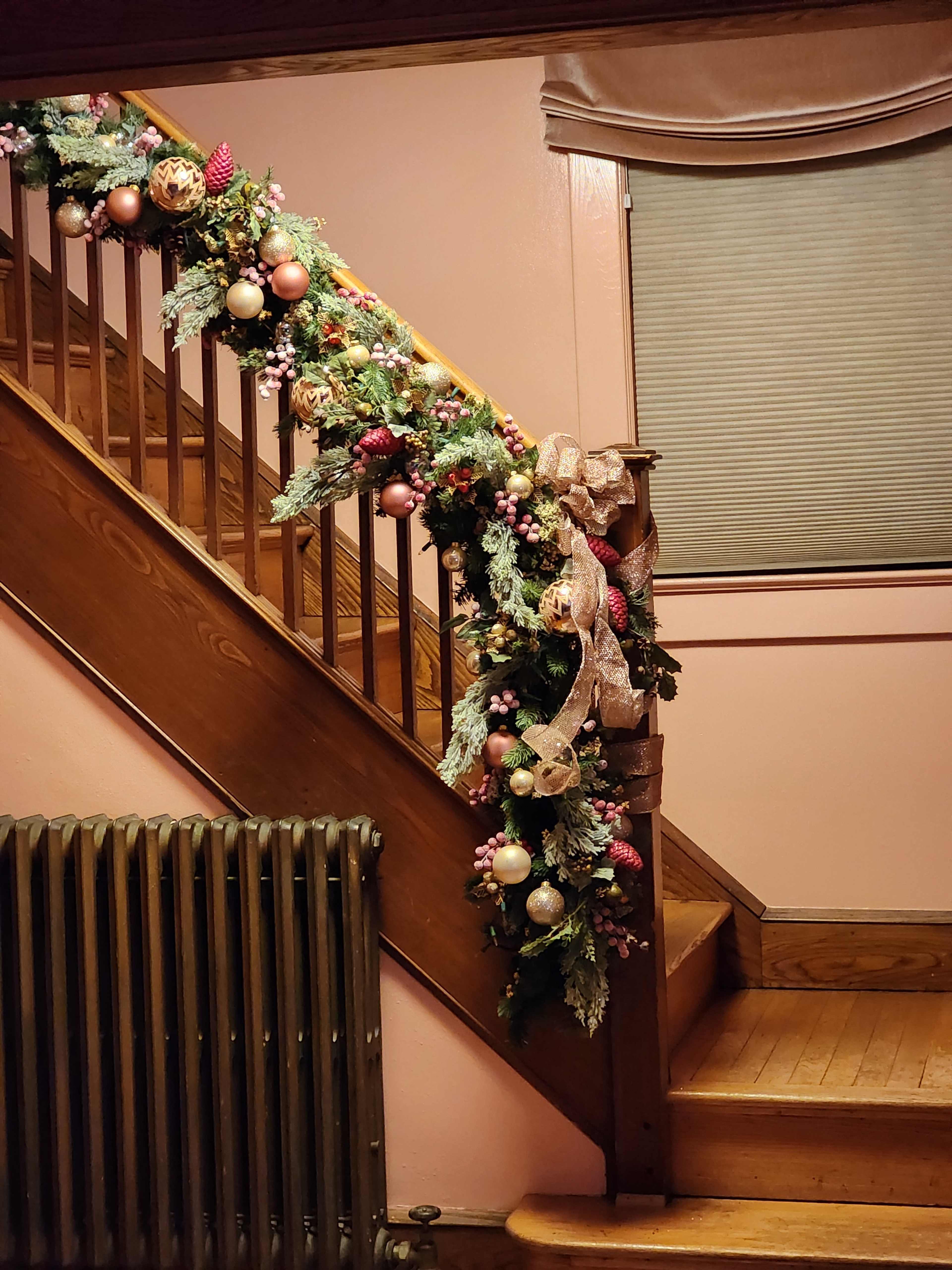 A decorated garland with pink ornaments and bows is draped over a wooden stair railing, beside a vintage radiator.