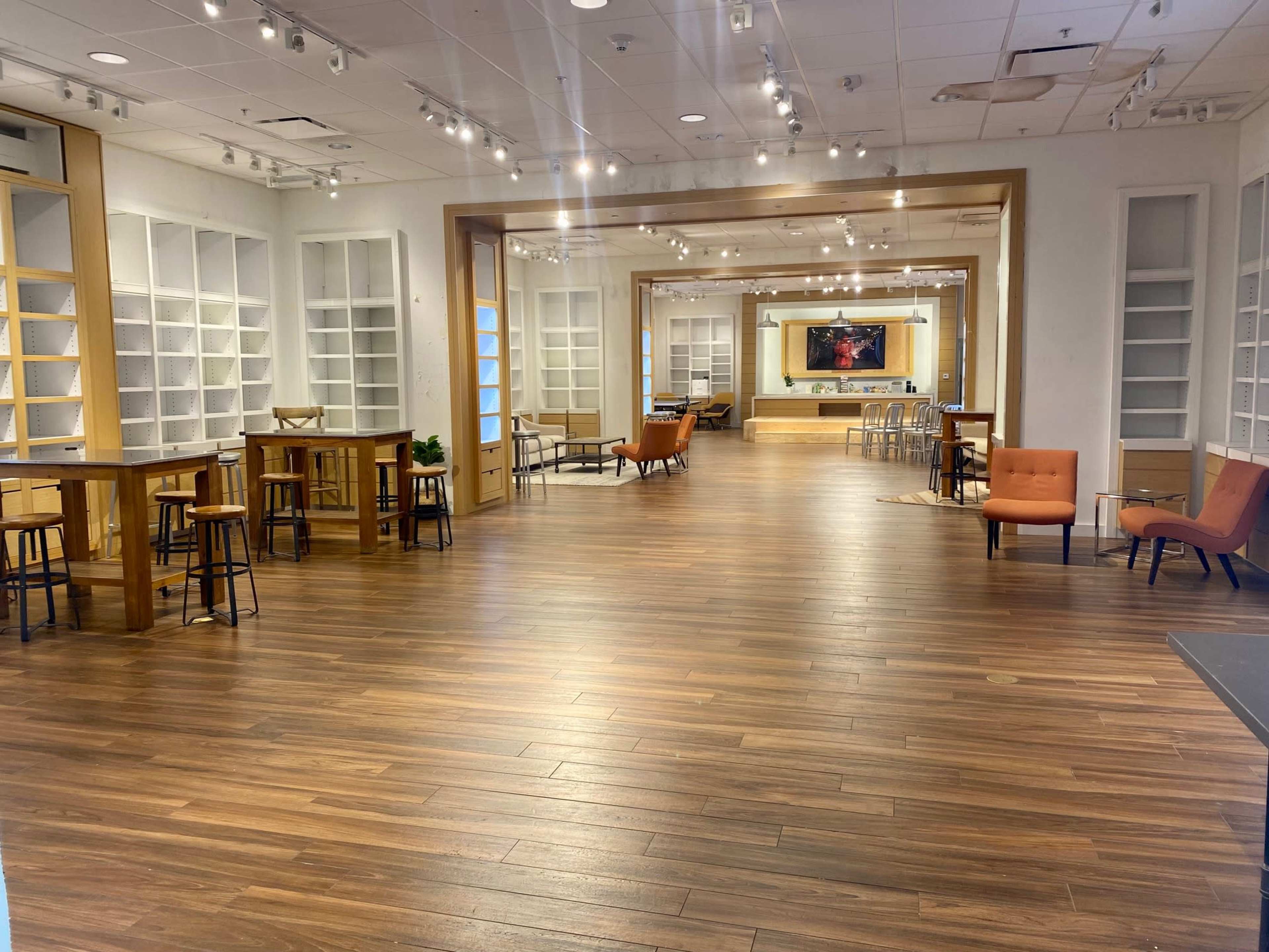 A spacious interior with wooden flooring, featuring tables, chairs, and a large mirror at the far end.