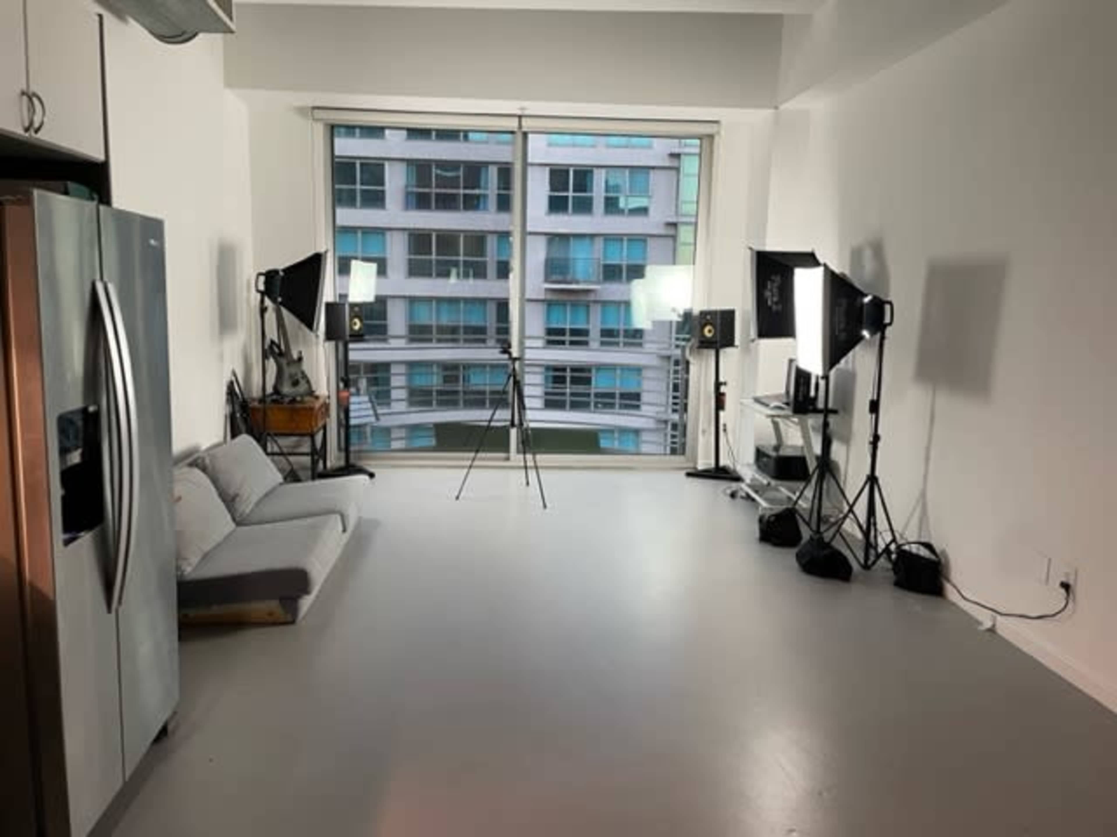 The image shows a spacious, well-lit room with a sofa, camera equipment, and studio lights set up near a large window.