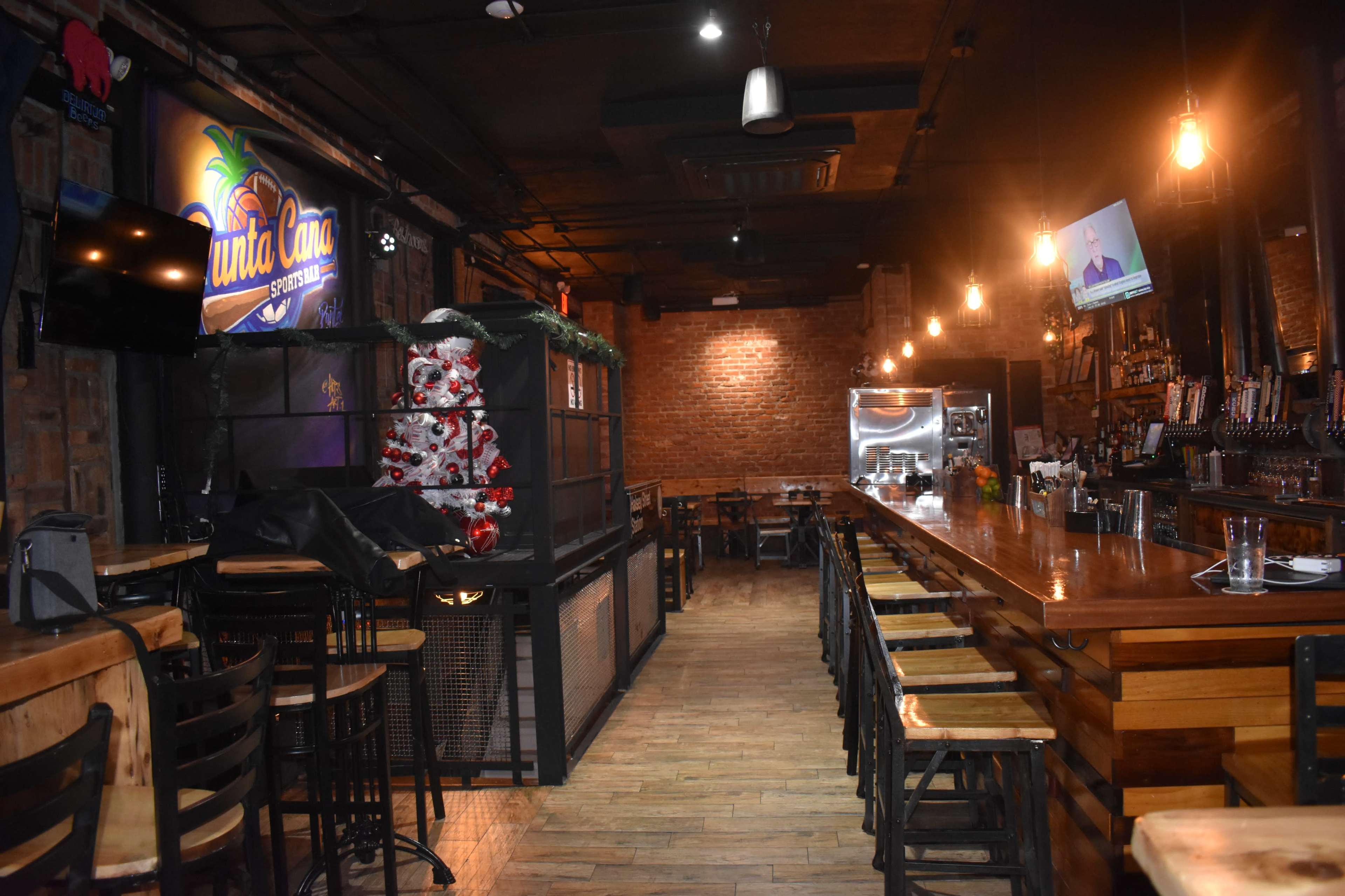 The image shows the interior of a dimly lit bar with wooden tables, a long counter lined with stools, and a brick wall in the background.