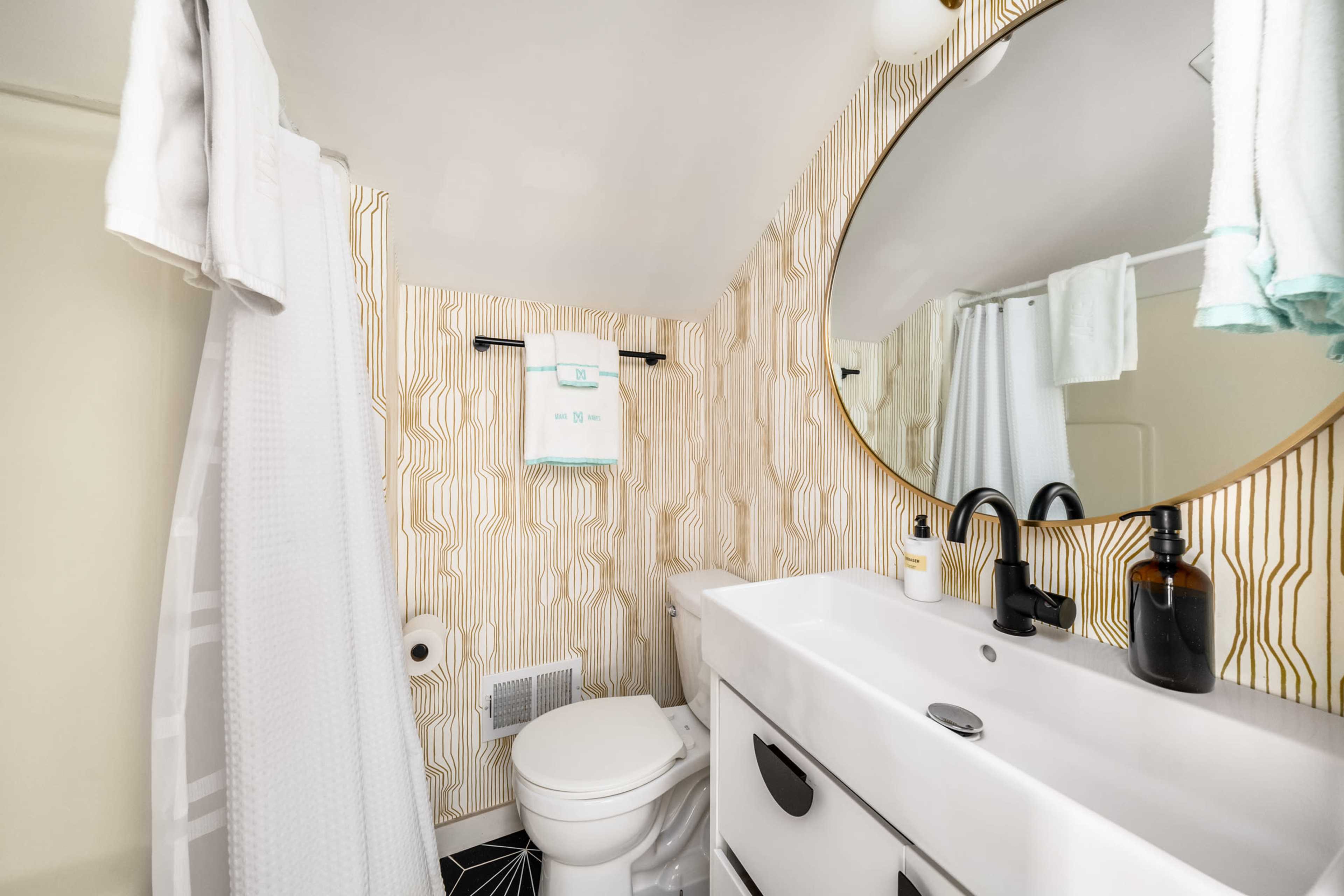 The image shows a modern bathroom featuring a round mirror above a white sink and toilet, with patterned wallpaper and a shower area.